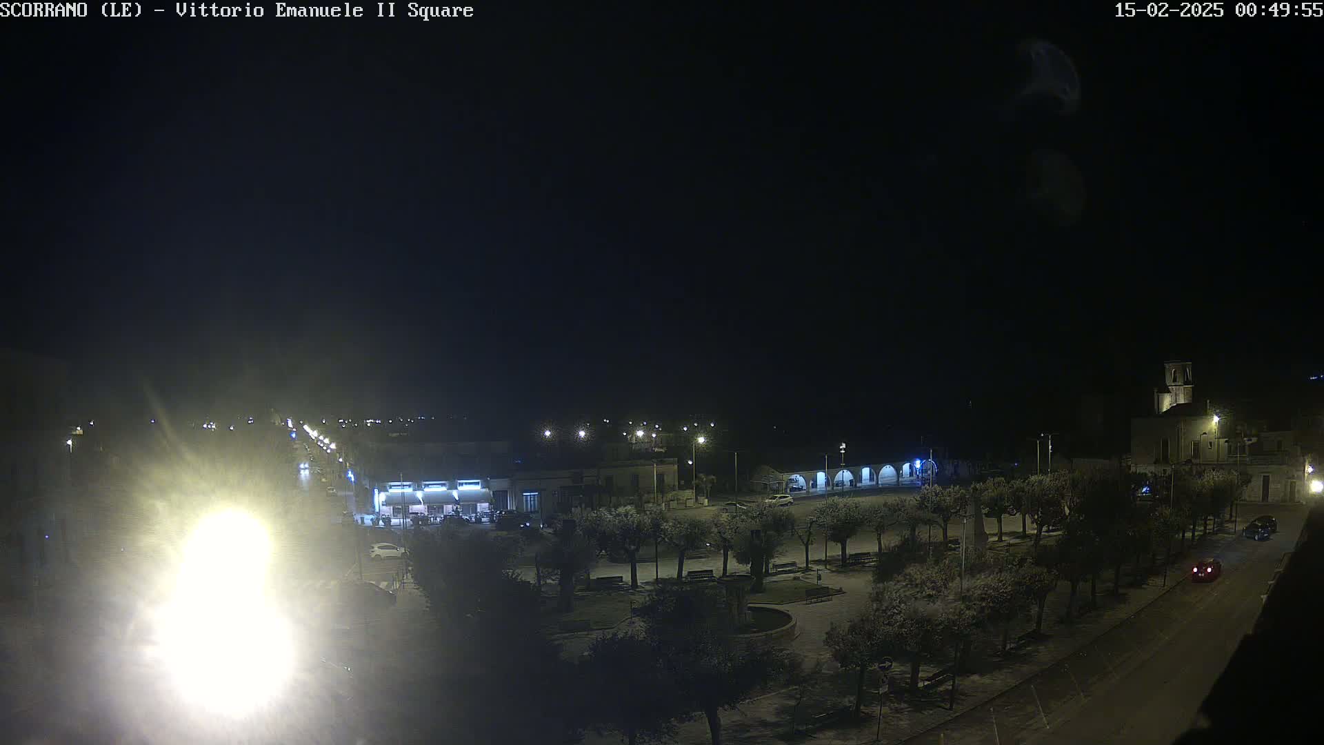 A nighttime view of a town square with a few cars on the road and trees lining the perimeter, under a clear, dark sky.