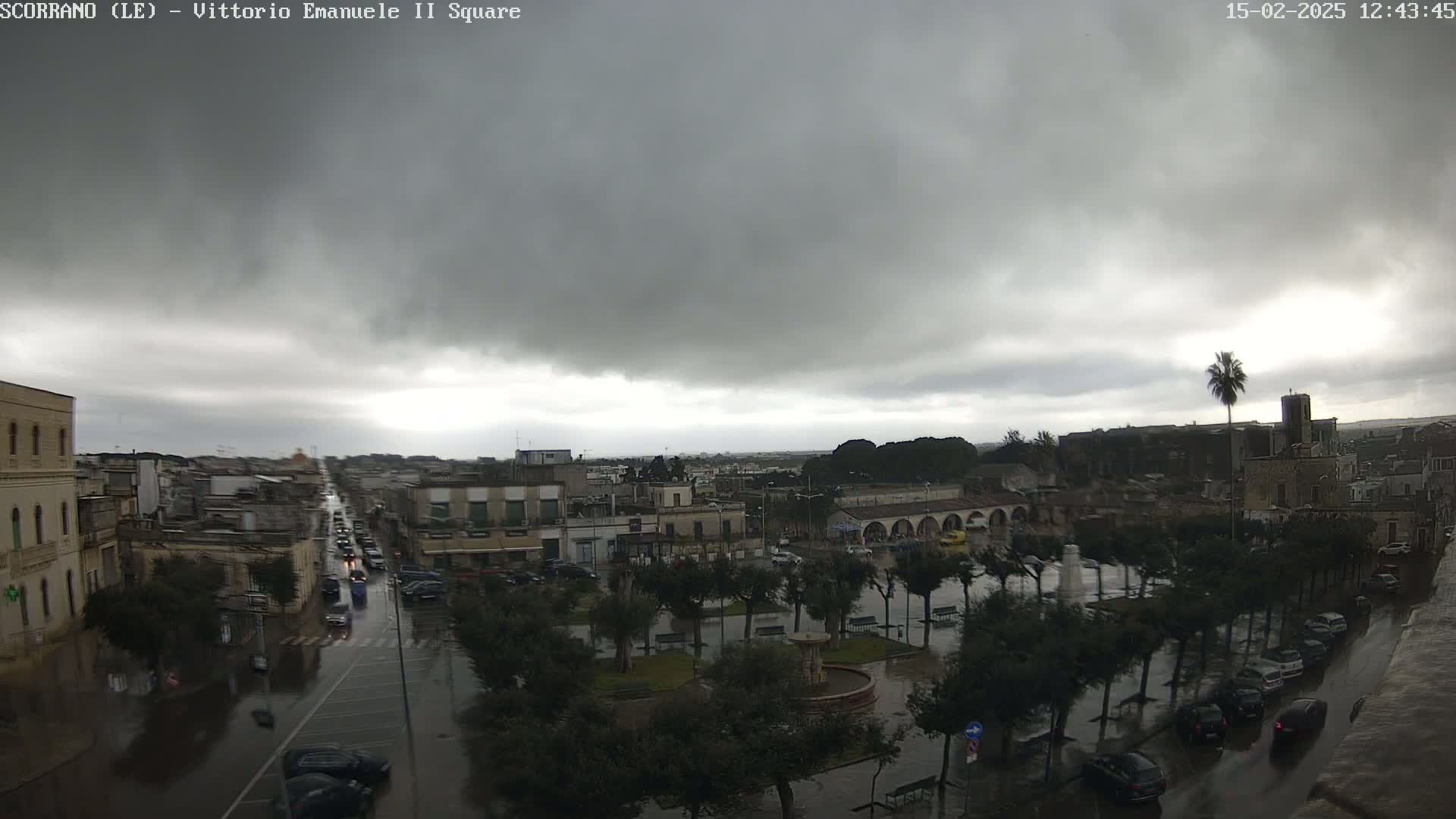 A rain-soaked town square, surrounded by buildings and trees under a dark, overcast sky.