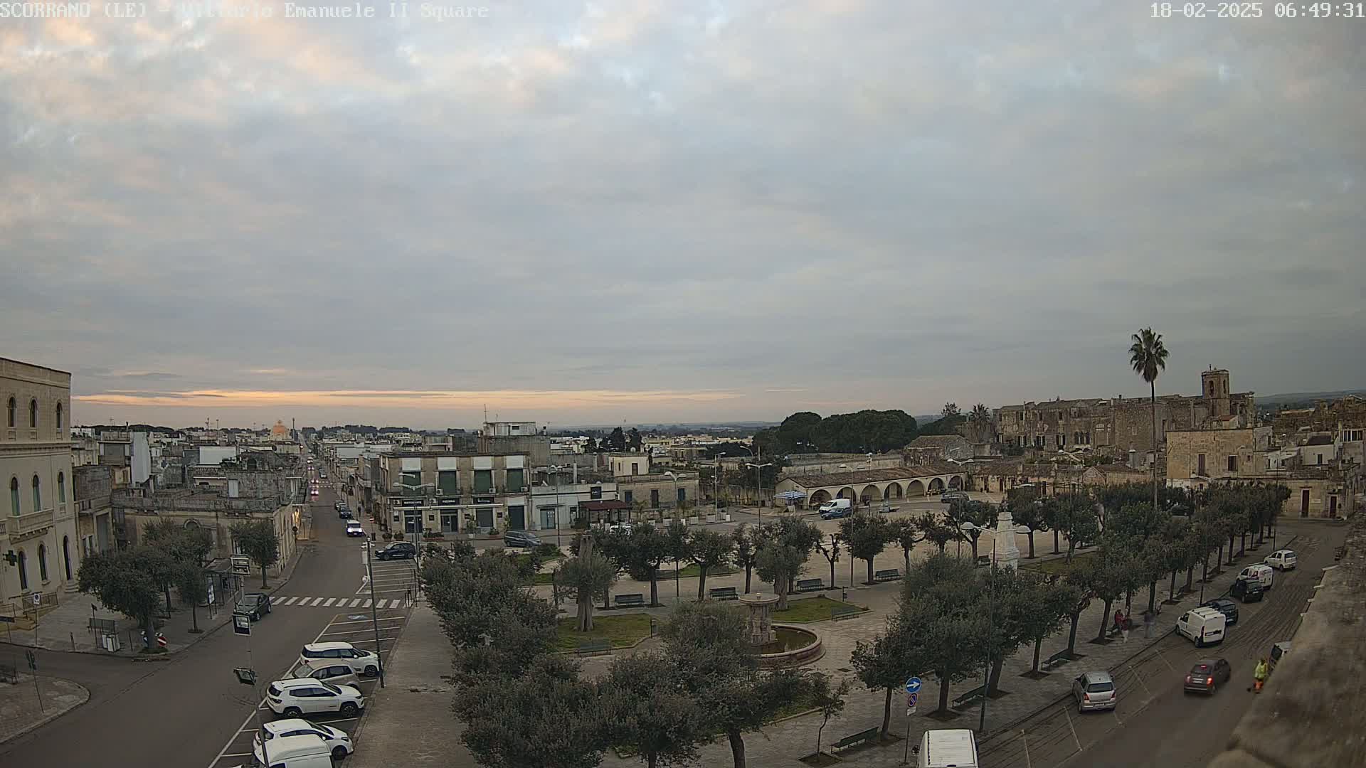 An overcast sunrise illuminates a town square with a fountain, surrounded by buildings and lined with trees, cars parked along the streets, and a few people visible.