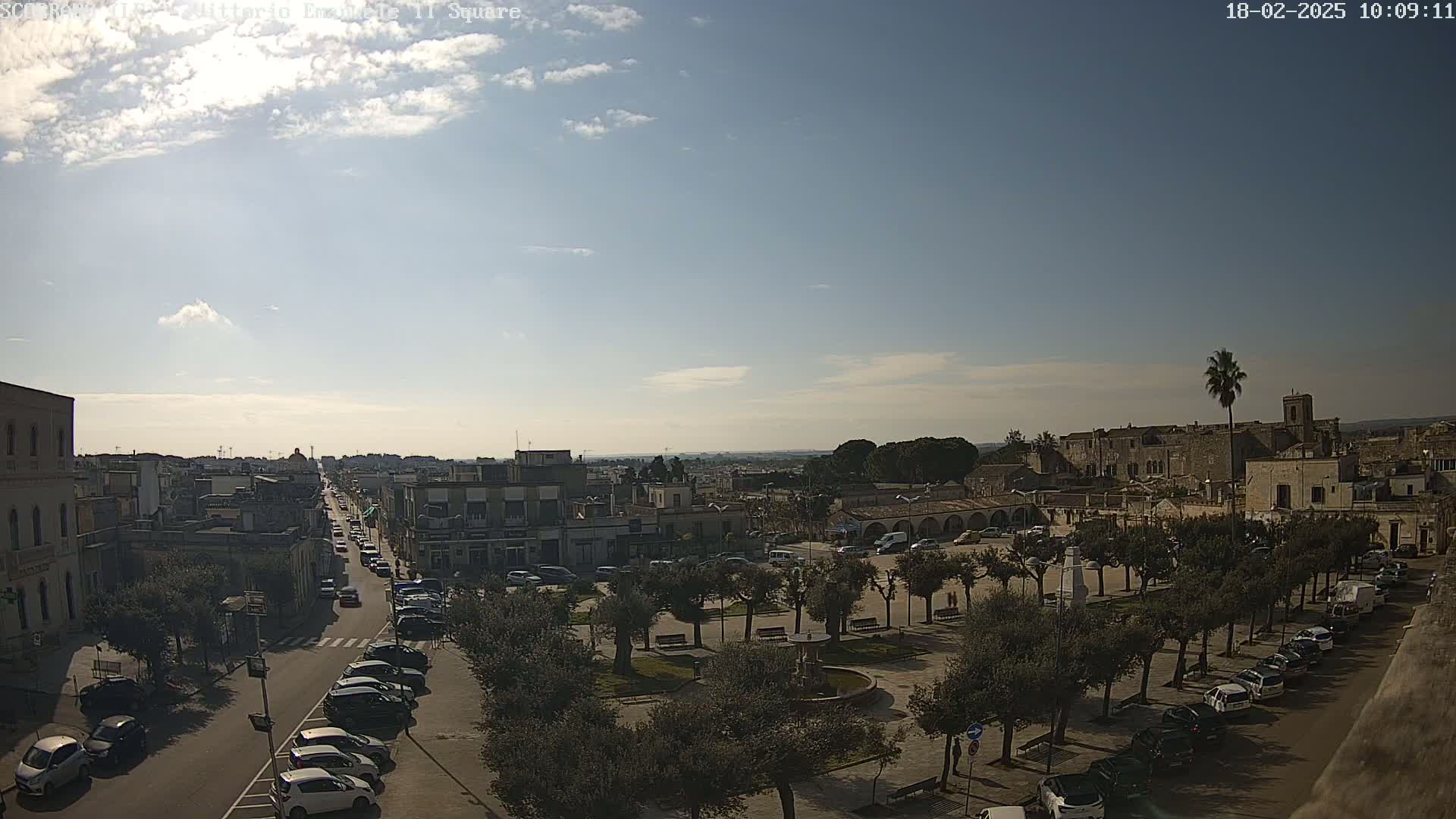 A mostly sunny day reveals a town square with a fountain, surrounded by parked cars and buildings, and a street leading to a distant horizon under a partly cloudy sky.