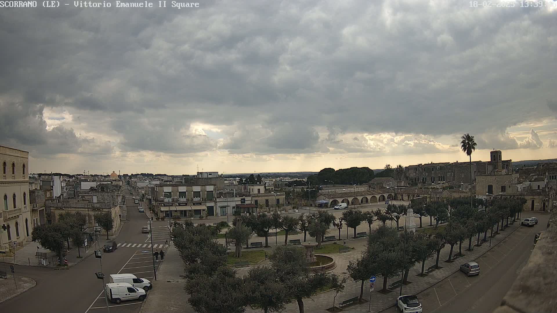 A town square with a fountain and numerous trees is visible under a cloudy sky.