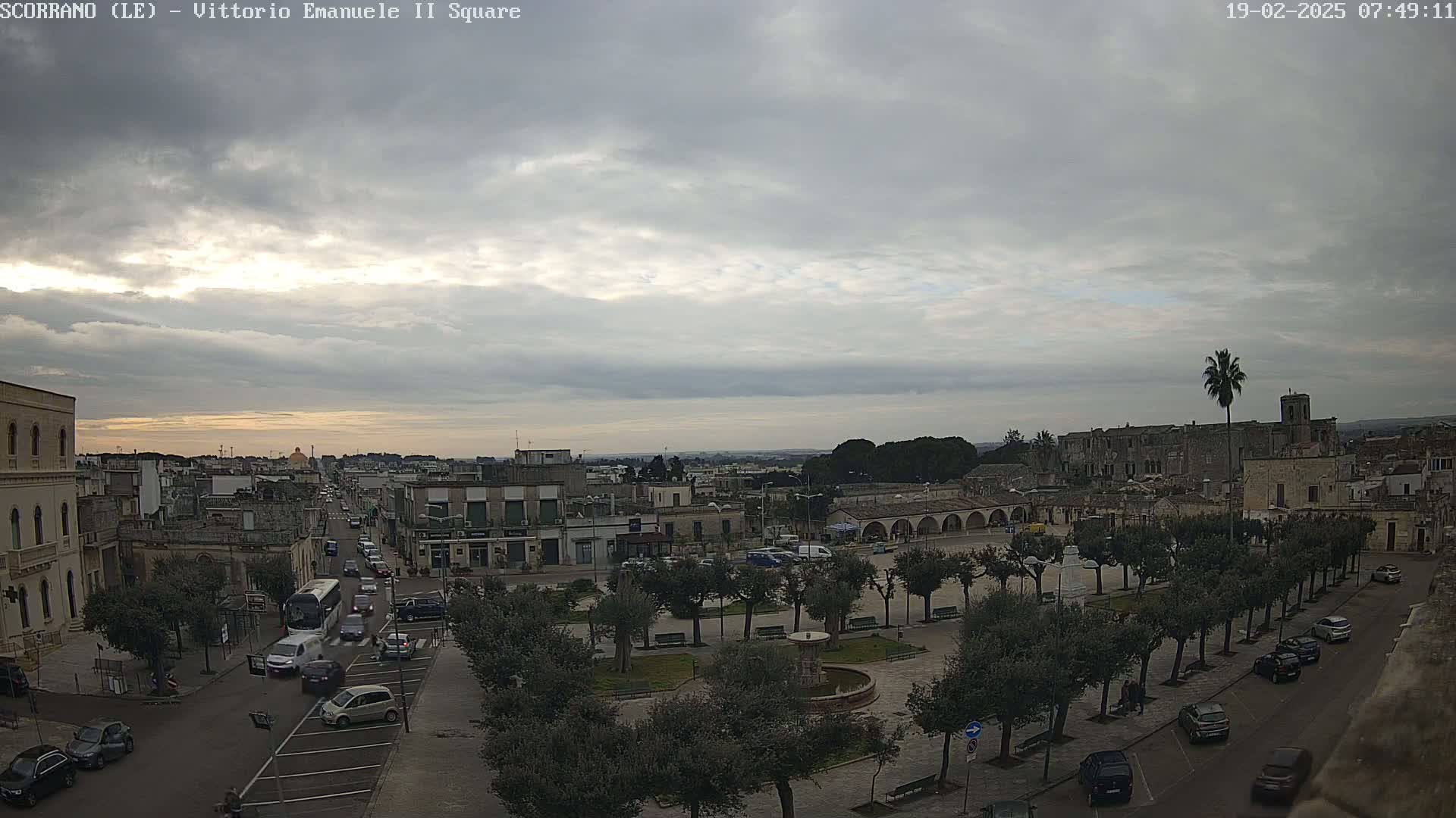 A mostly cloudy sky hangs over a town square with several parked cars and a few pedestrians, where many olive trees line the perimeter of the square.