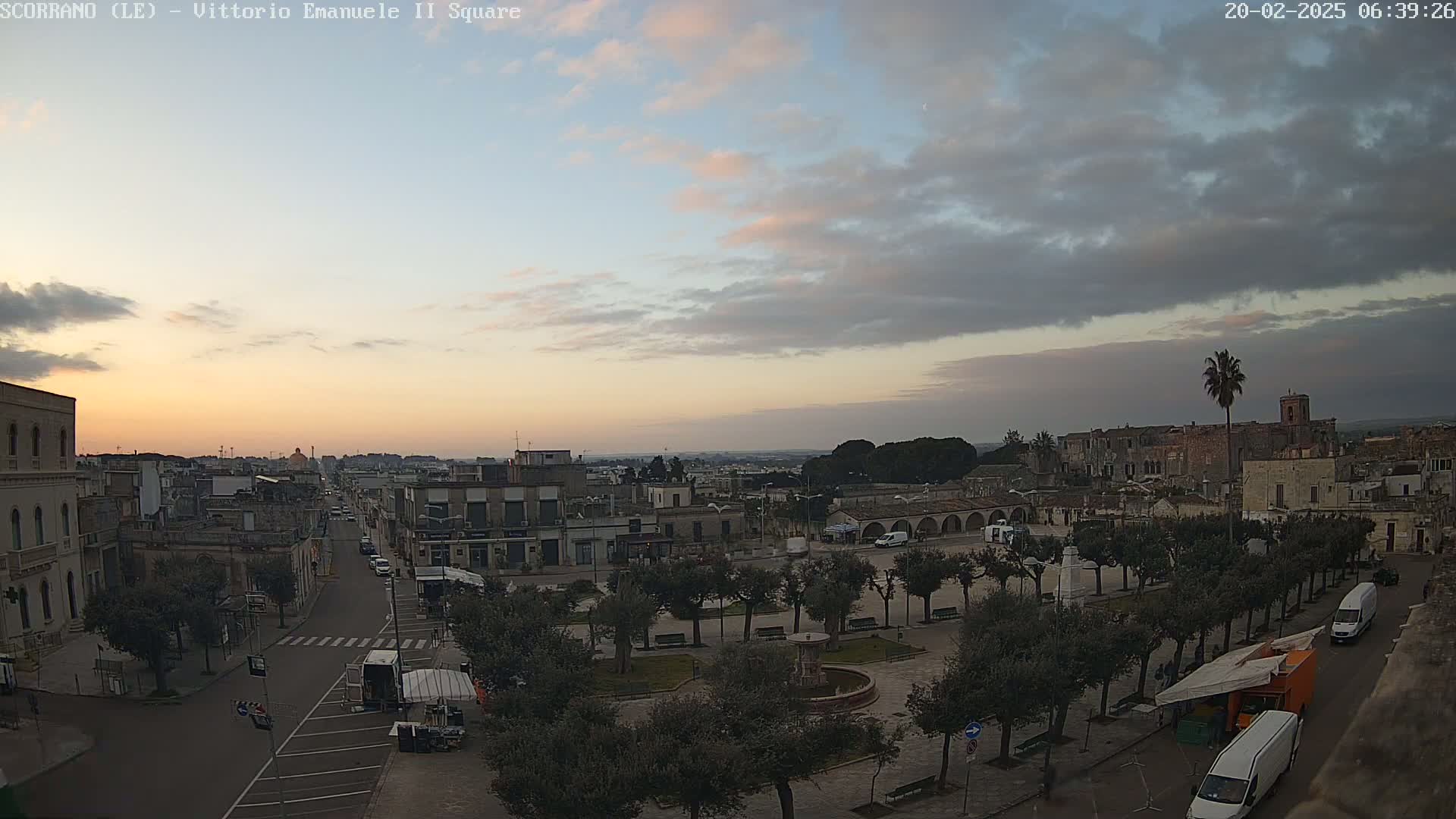 A partly cloudy sunrise illuminates a town square with a fountain, surrounded by buildings and trees, with vehicles parked along the street.