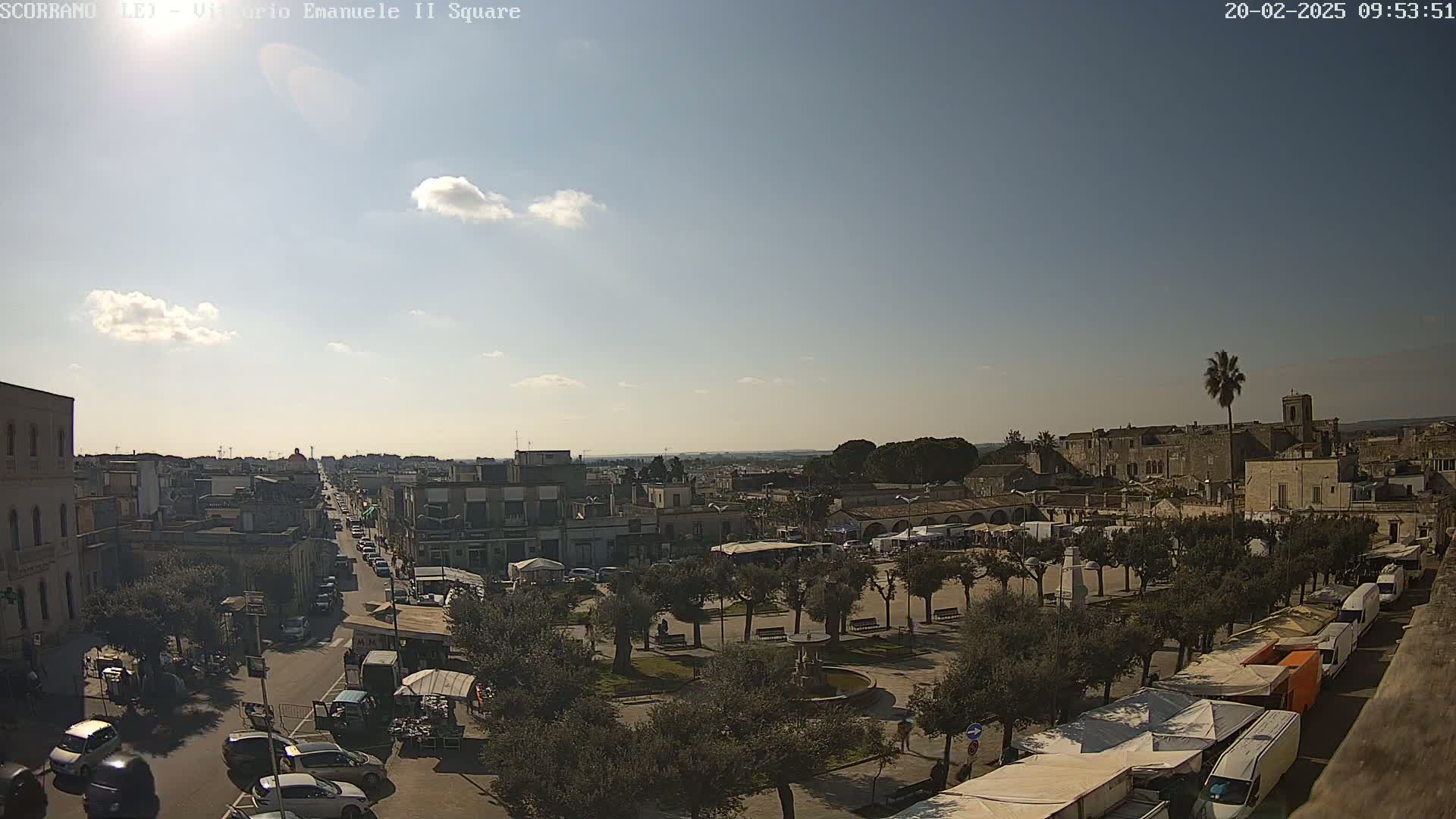 A sunny day reveals a town square with a market, surrounded by buildings and a street with parked cars.