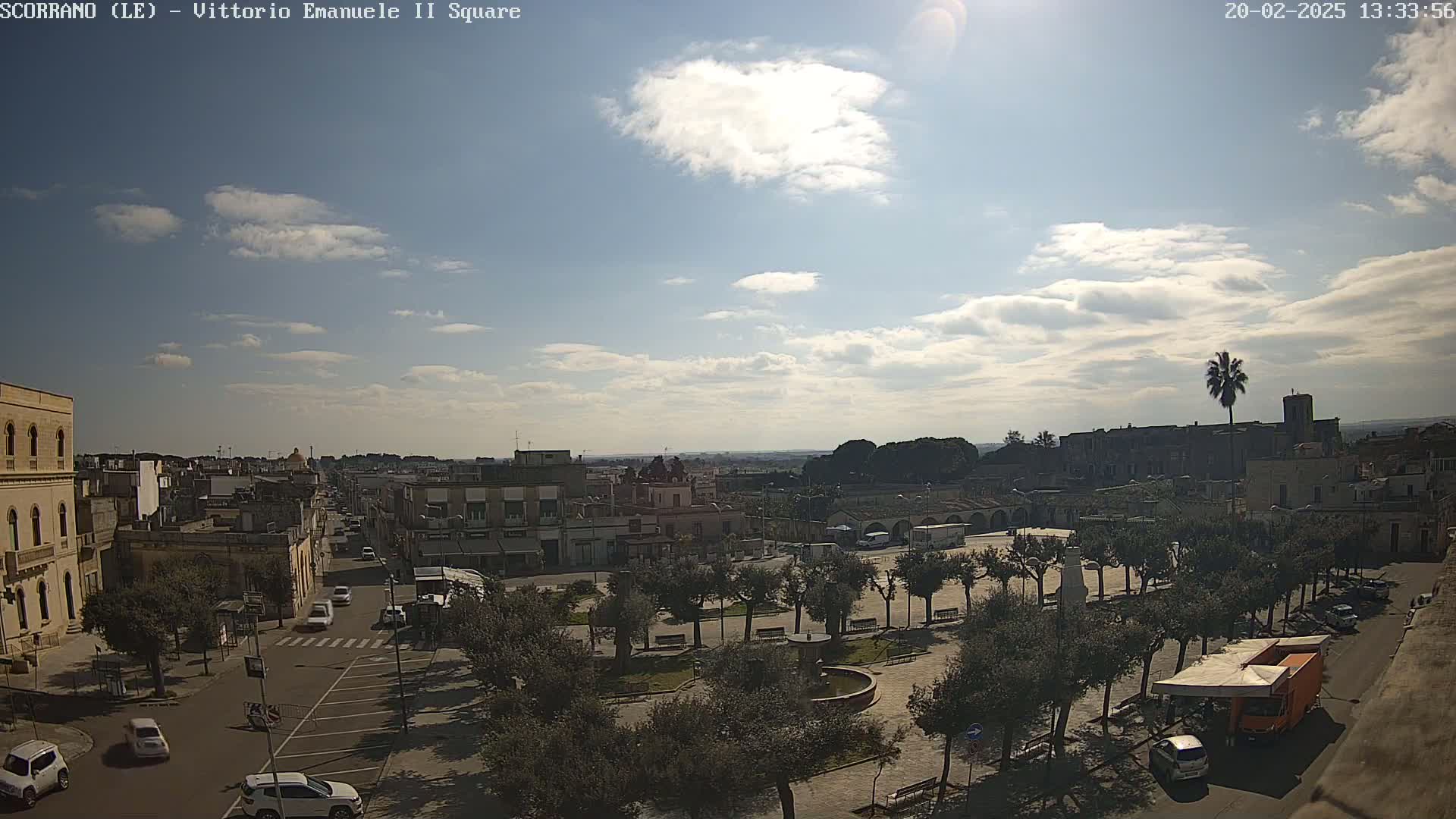 A partly cloudy daytime view of a town square with trees, buildings, and vehicles, under a mostly sunny sky.