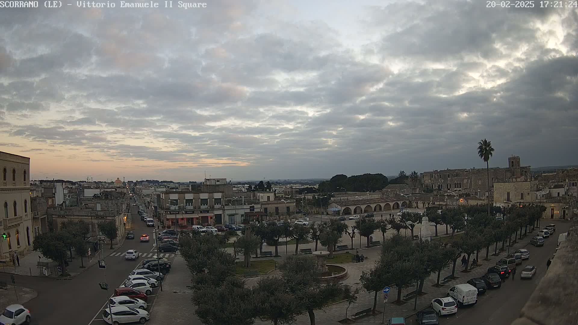 A town square with parked cars and trees is seen under a mostly cloudy sky at dusk.