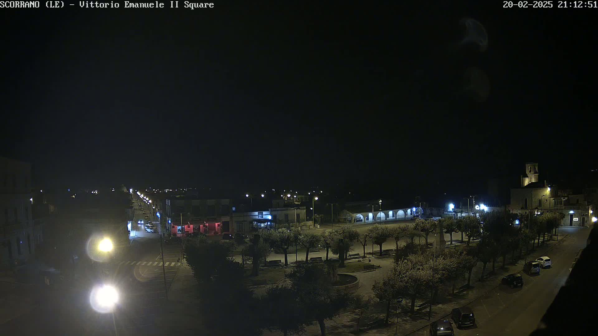 A nighttime, overhead view of a town square with sparsely parked cars and trees lining the perimeter, under a clear, dark sky.