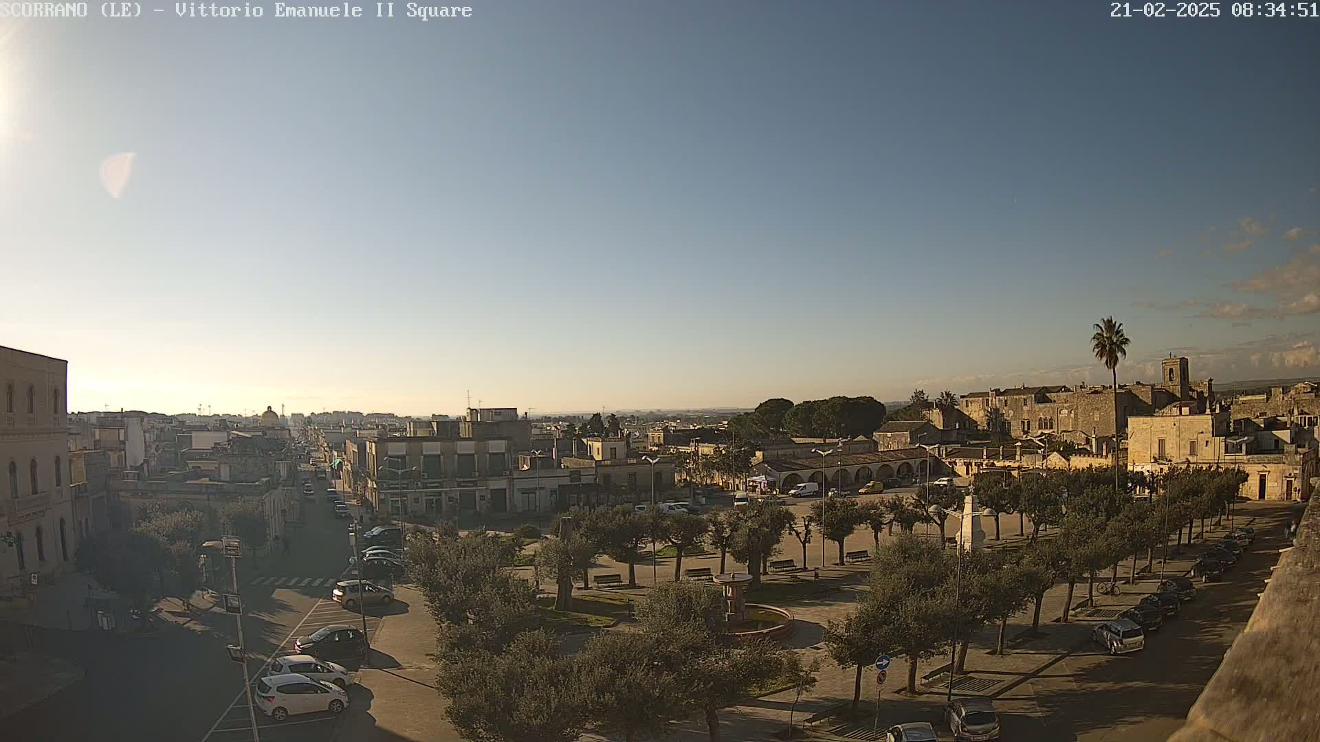 A clear, sunny day reveals a town square surrounded by low buildings and trees, with parked cars lining the streets and a view of a larger town or city in the background.
