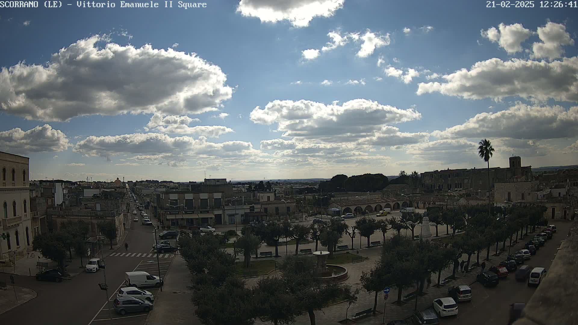 A partly cloudy day overlooks a town square with parked cars and trees, a street leading into the distance, and buildings surrounding the area.