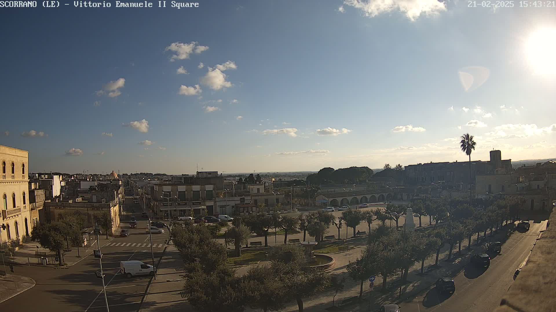 A sunny day reveals a town square with parked cars and a few trees, surrounded by buildings and a wider cityscape under a partly cloudy sky.