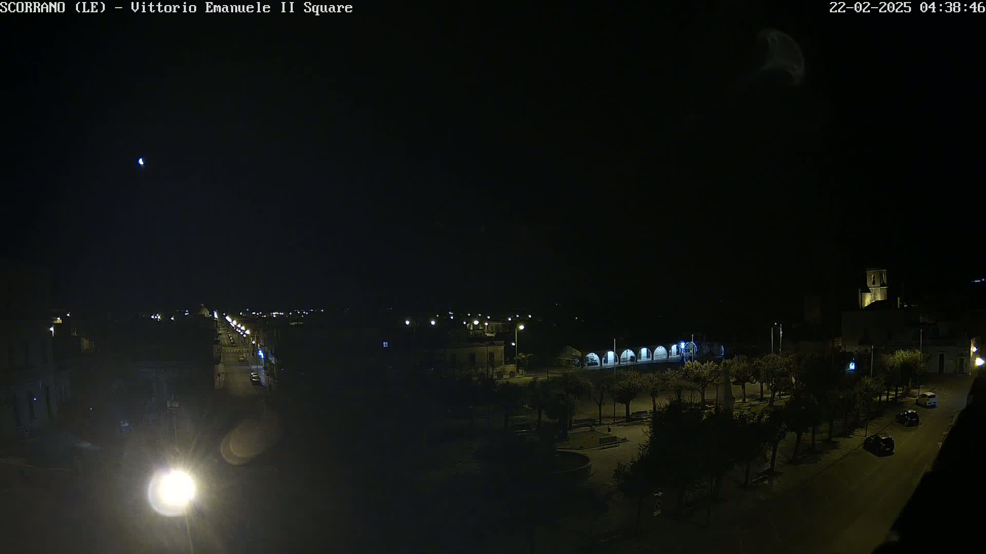 A nighttime, overhead view of a town square with a few cars on the road, a crescent moon visible in the dark sky.