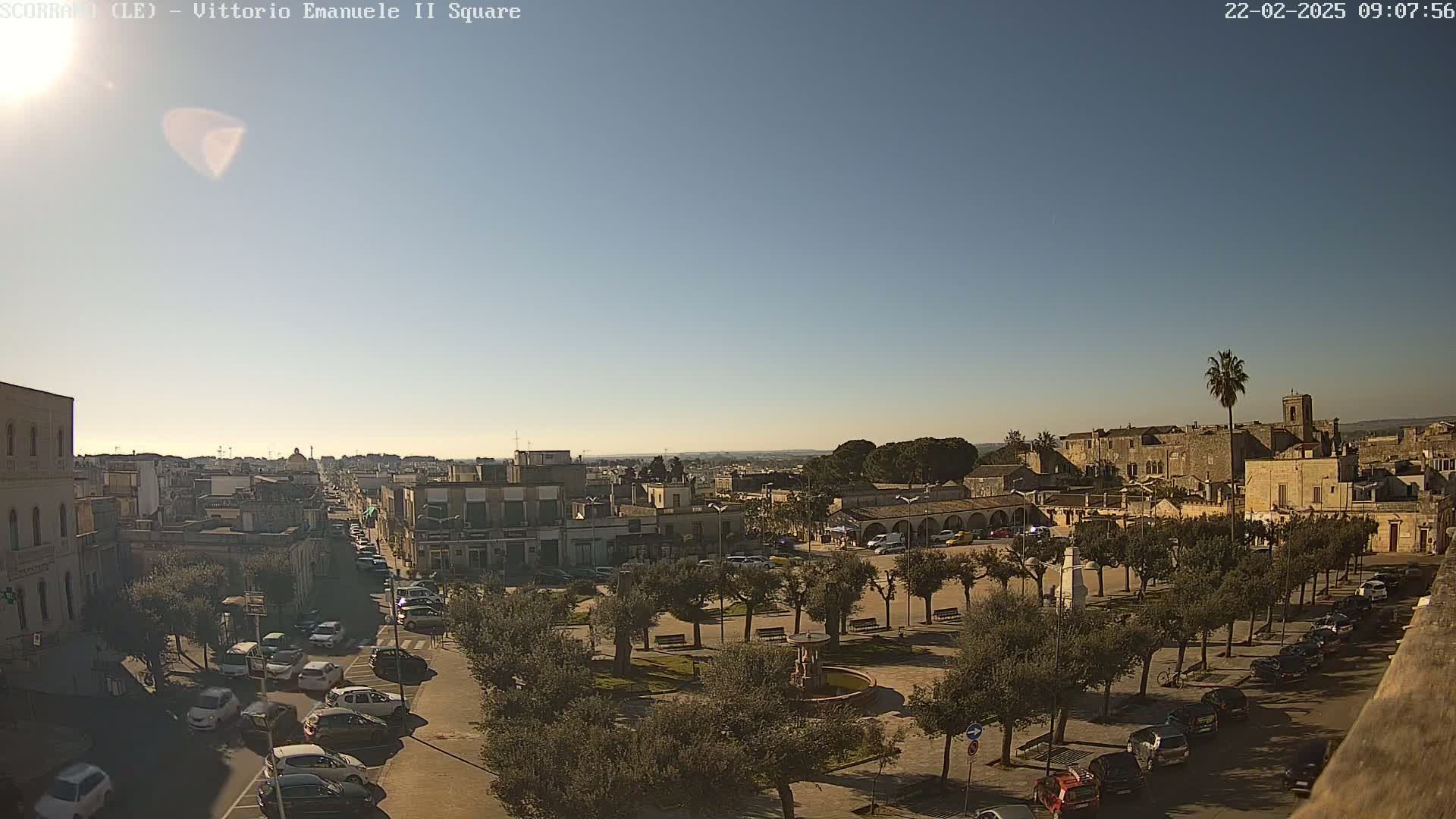 A sunny, clear day reveals a high-angle view of a town square surrounded by buildings and parked cars, with a fountain at its center and trees lining the perimeter.
