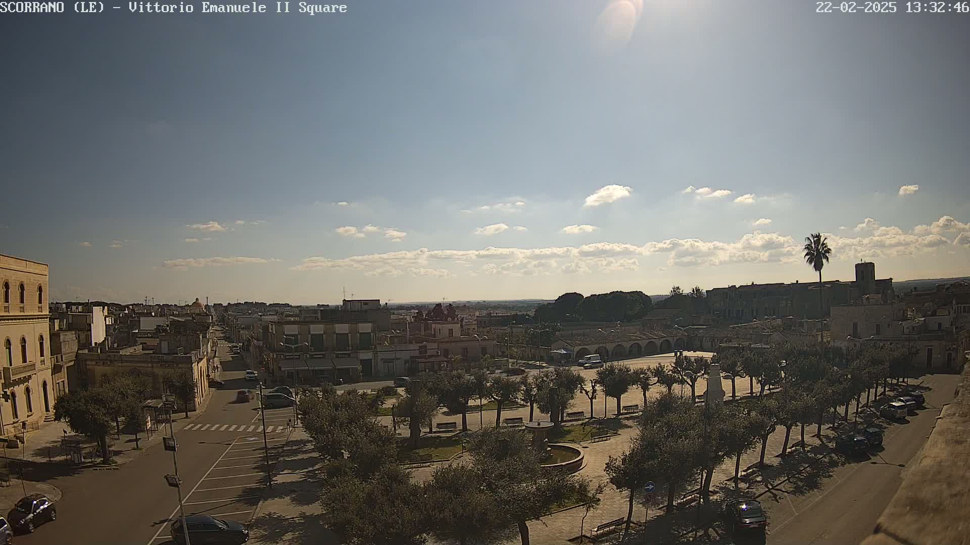 A sunny day reveals a town square with numerous trees, parked cars lining a road, and buildings extending into the distance under a partly cloudy sky.