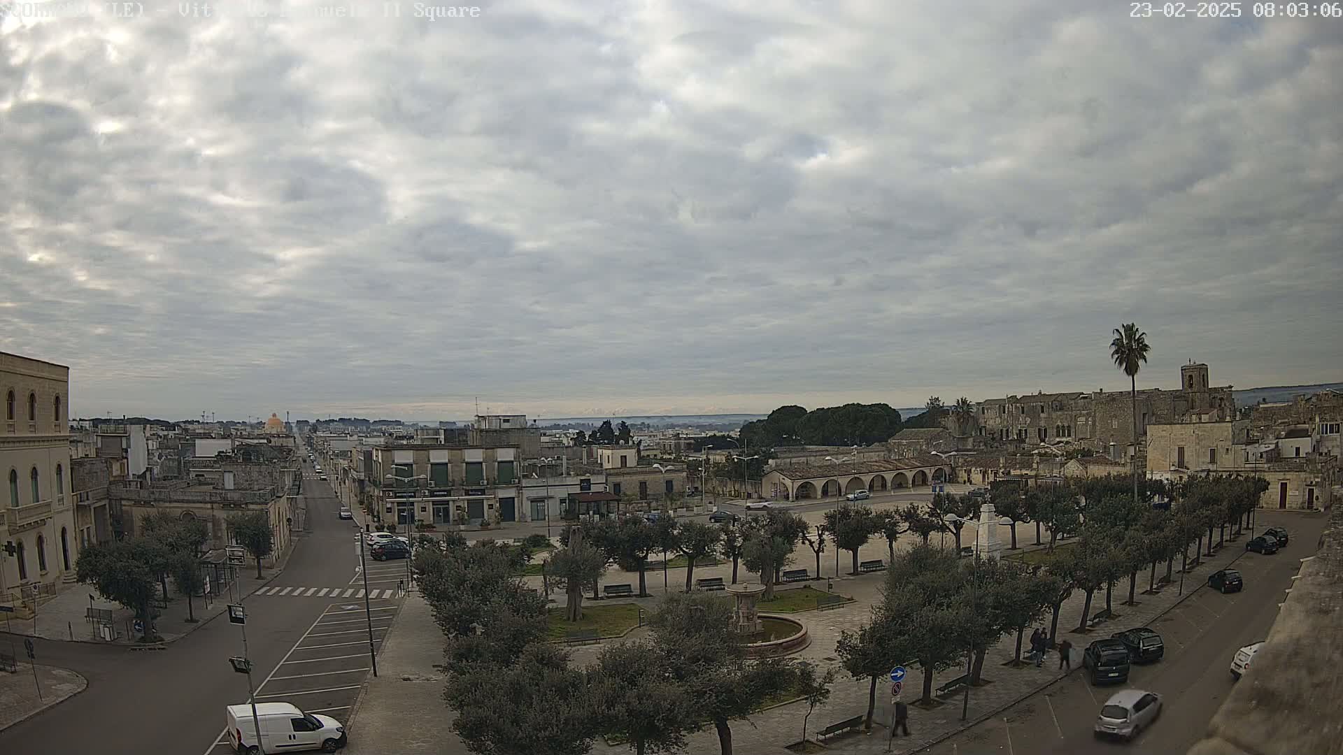 An overcast day reveals a town square with a fountain, surrounded by buildings and trees, a street leading to a distant cityscape under a cloudy sky.