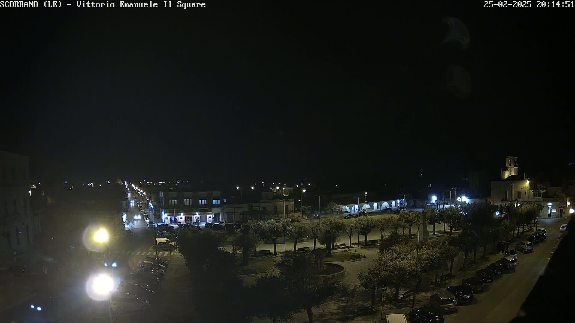 A nighttime aerial view of a town square with parked cars lining the streets and illuminated trees in the center.