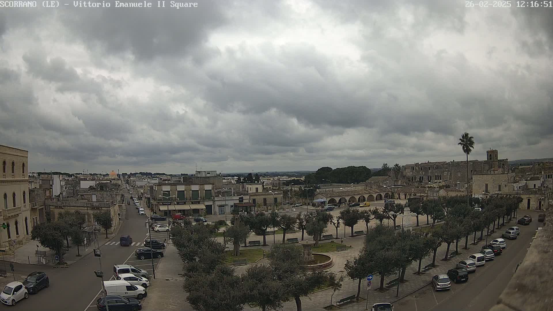 A town square with parked cars and a fountain is viewed from above under an overcast sky.
