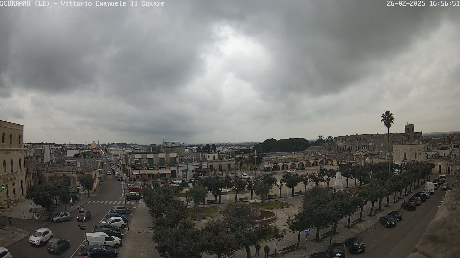 A town square with parked cars and a central fountain is visible under an overcast sky.