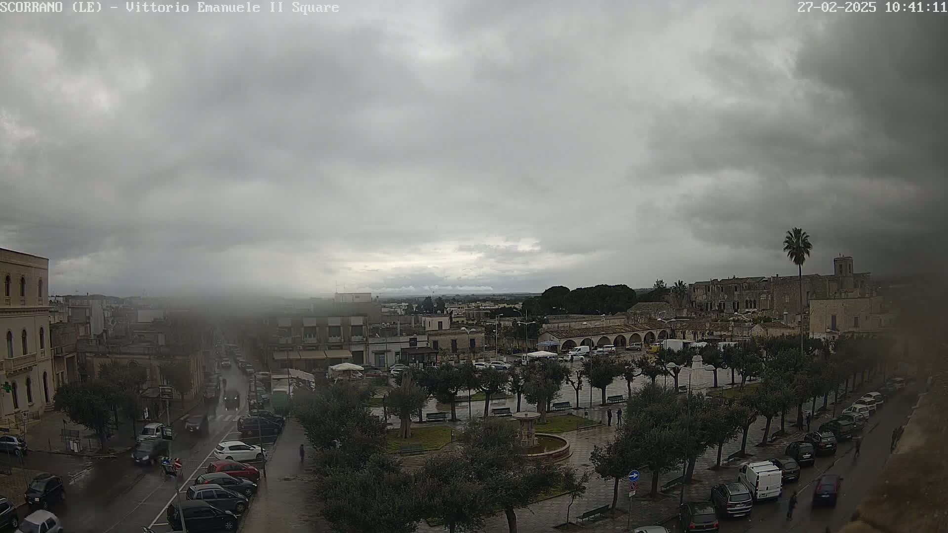 A town square with a fountain and surrounding buildings is seen under a cloudy, overcast sky, with rain falling lightly on the wet streets and parked cars.