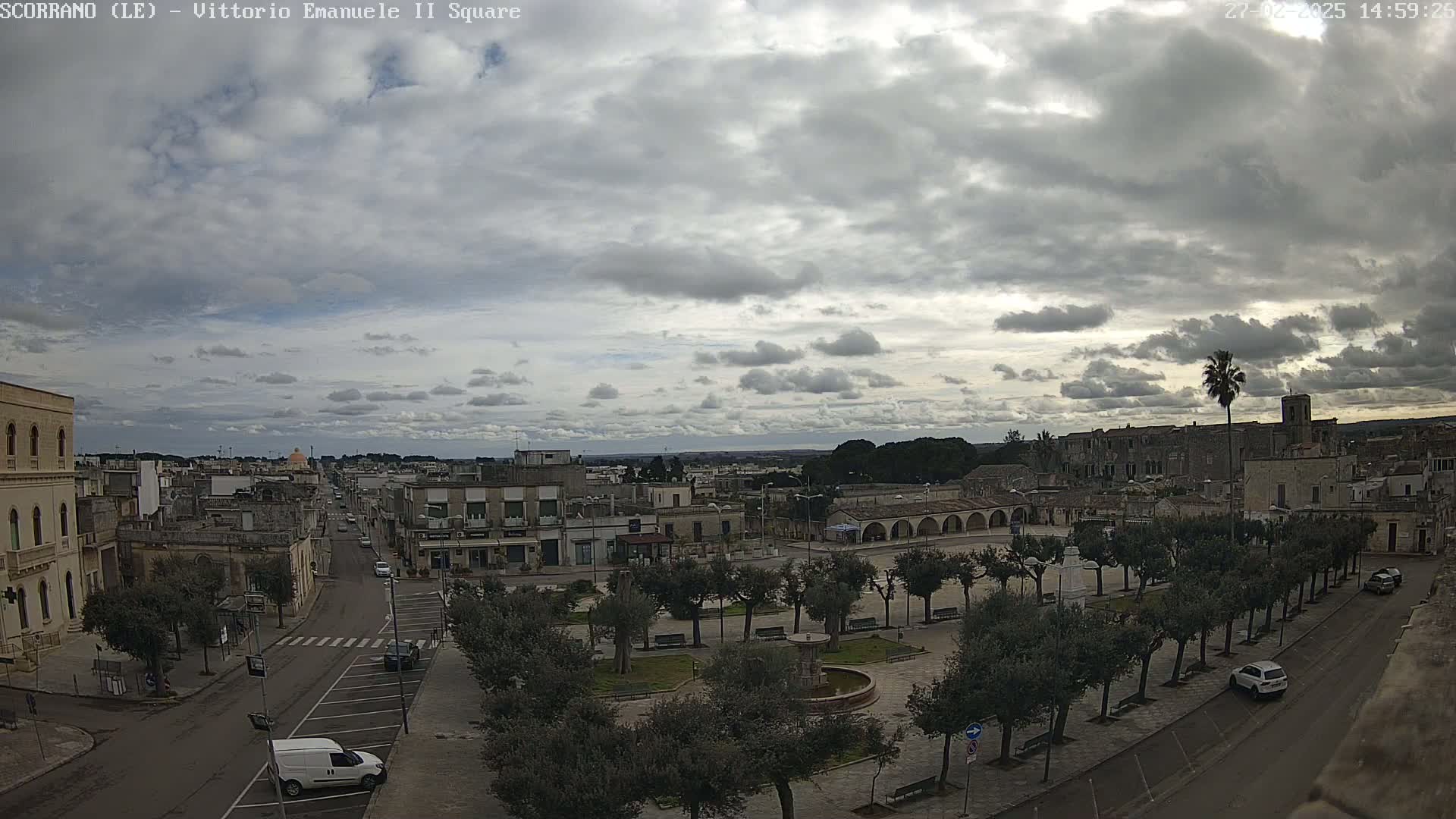An overcast sky hangs over a town square with a fountain, lined with trees and a few parked cars, and a street leading to buildings and more of the town beyond.