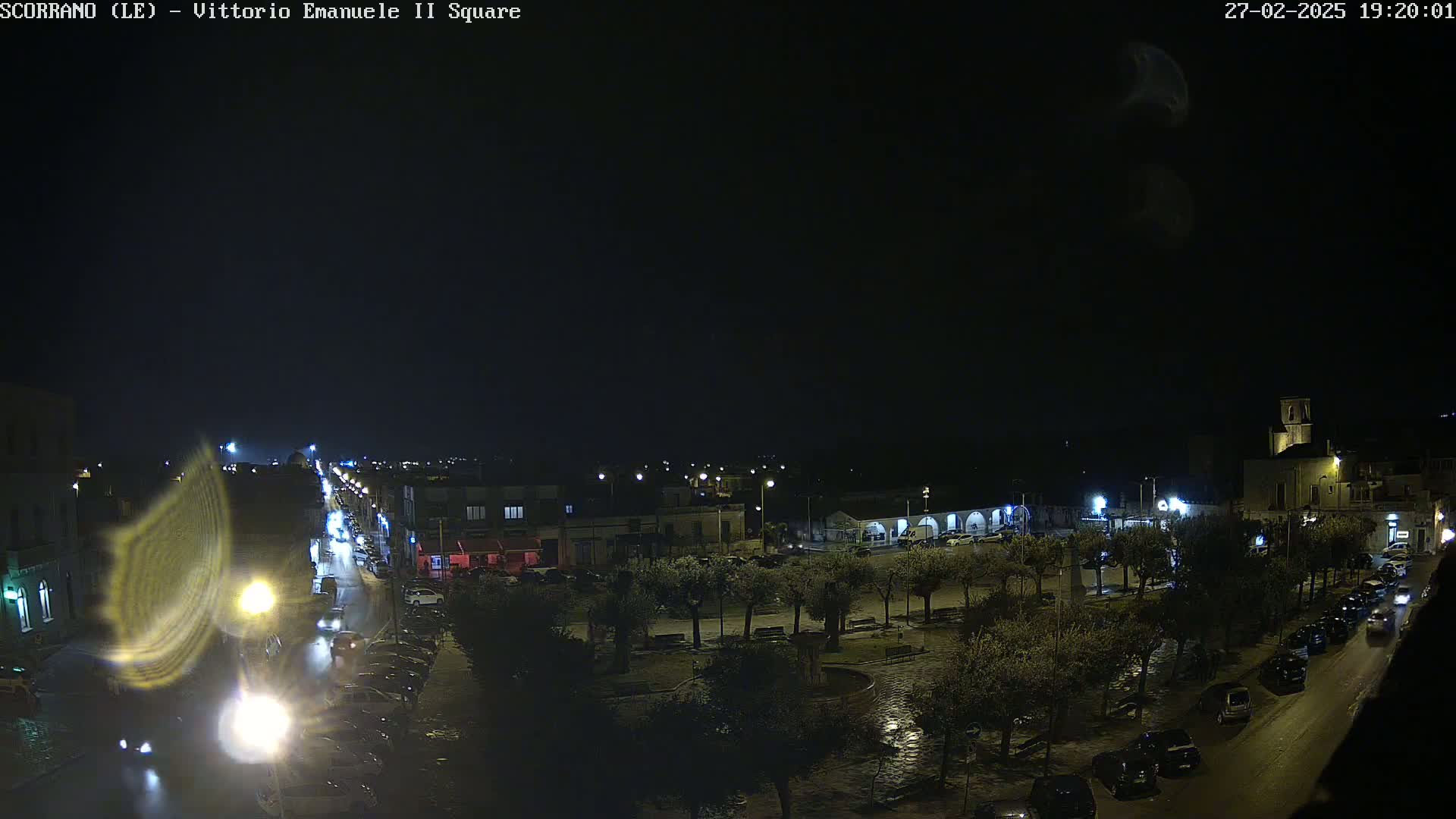 A nighttime, wet view of a town square with cars parked along the streets and trees lining the square.