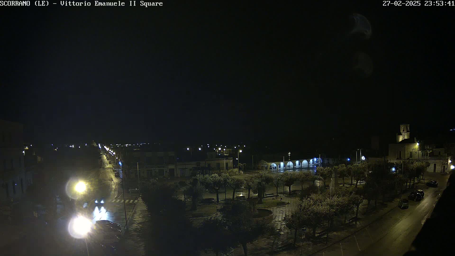 A nighttime view of a rain-slicked town square with streetlights illuminating buildings, trees, and a few vehicles.