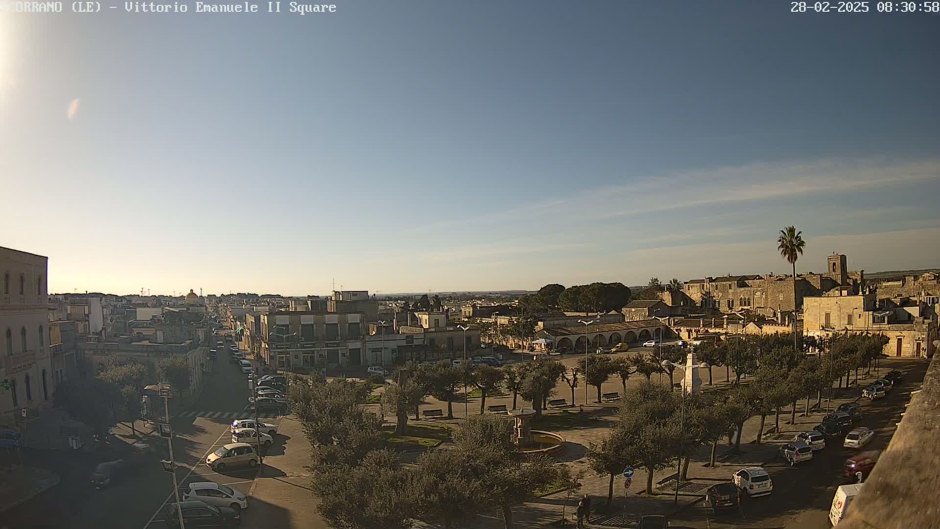 A sunny day reveals a town square with parked cars, lined with trees, and buildings in the background.