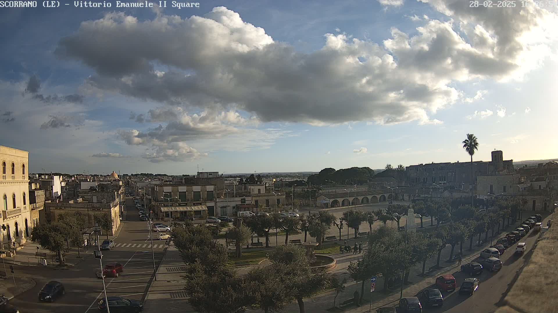 A partly cloudy sky overlooks a town square with parked cars and buildings, some trees lining the streets.