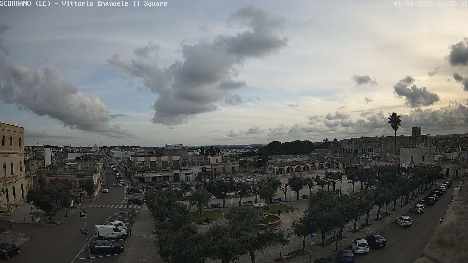 A partly cloudy sky overlooks a town square with parked cars and trees lining the streets, and buildings extending into the distance.