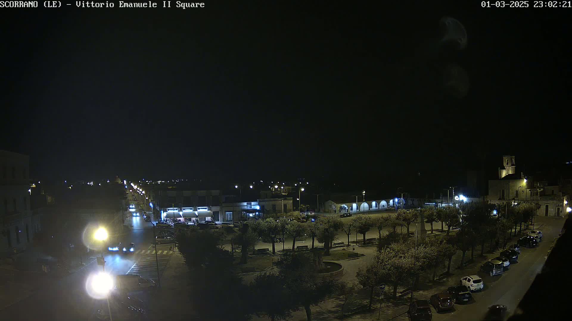 A nighttime, overhead view of a town square with cars parked along the sides and a few cars driving on the road, under a clear, dark sky.