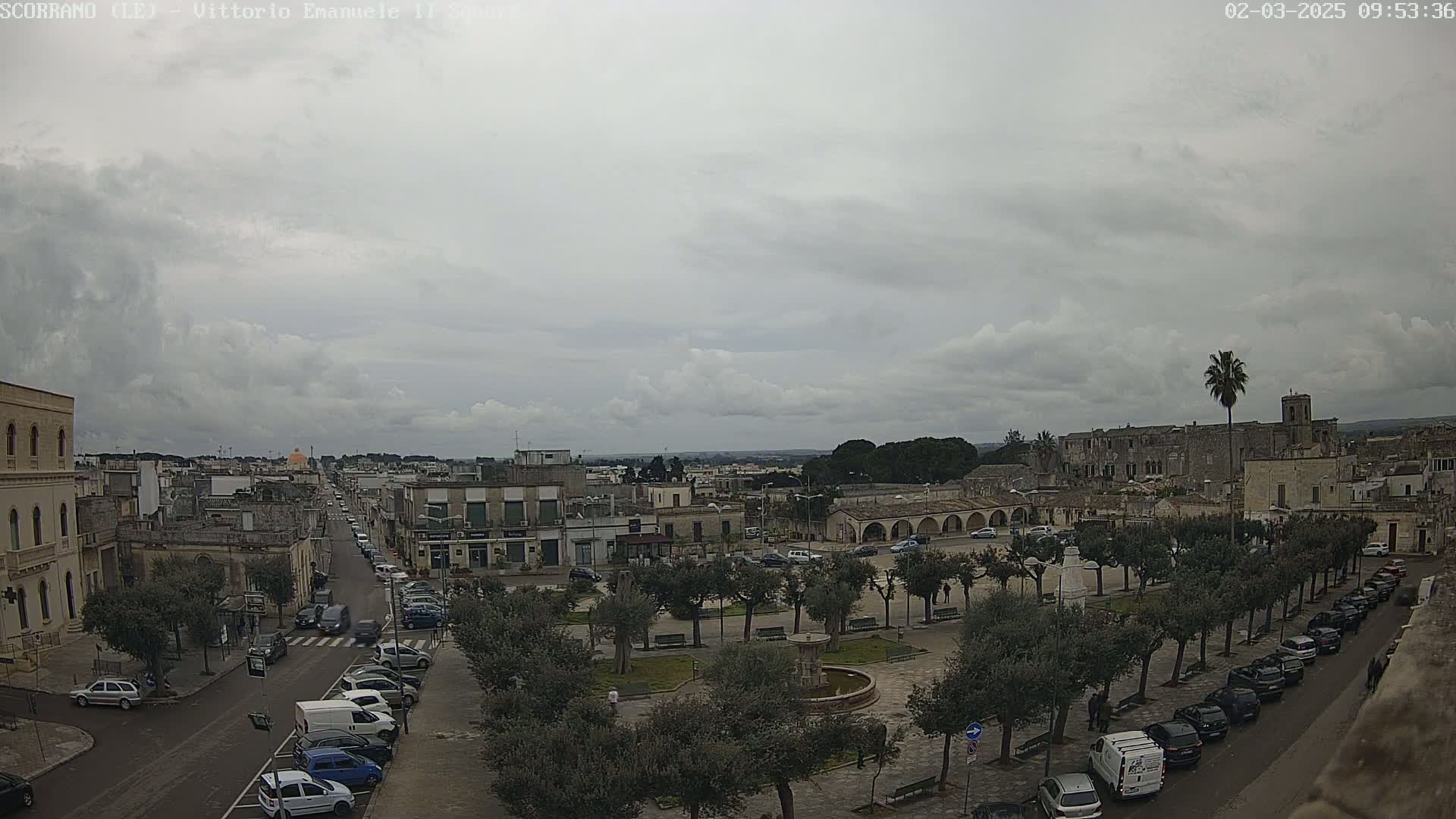 A mostly cloudy day reveals a town square with parked cars lining a street, several buildings, and a fountain, all surrounded by trees.