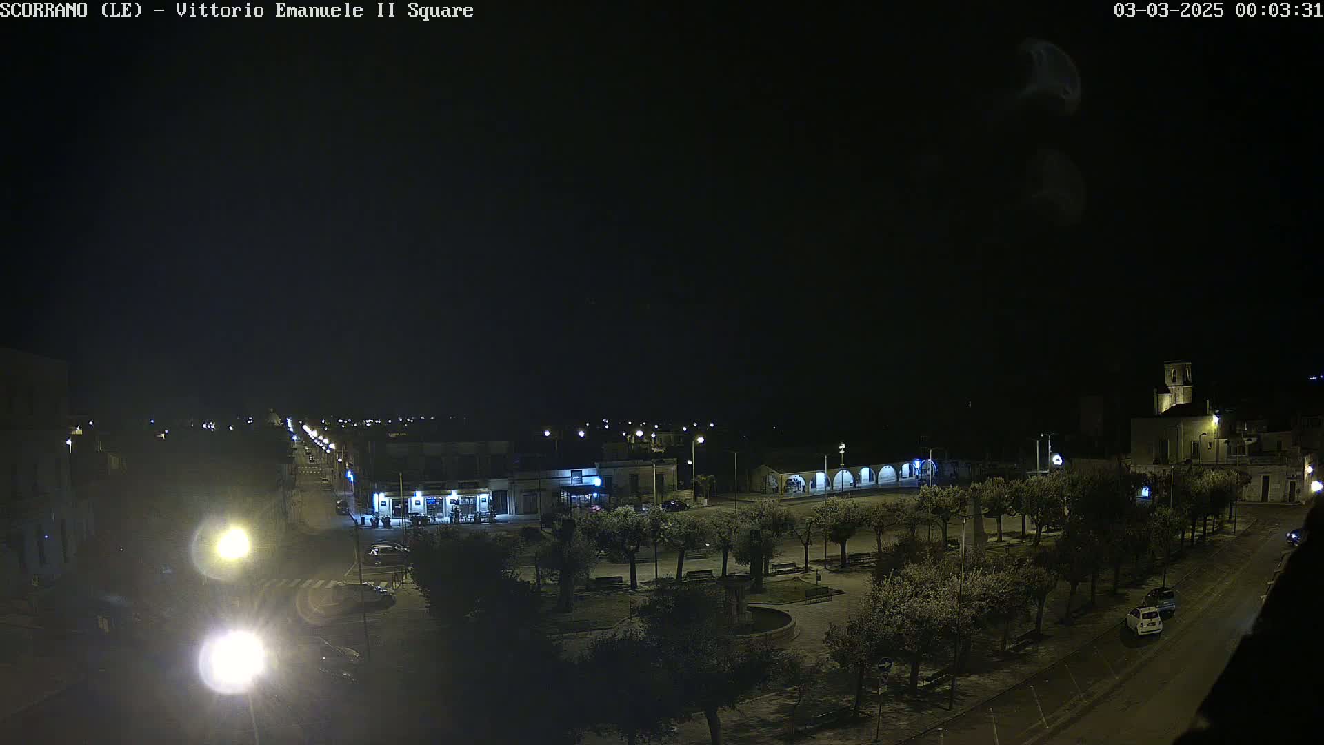 A nighttime, dimly lit town square with sparsely parked cars and trees under a dark, clear sky.