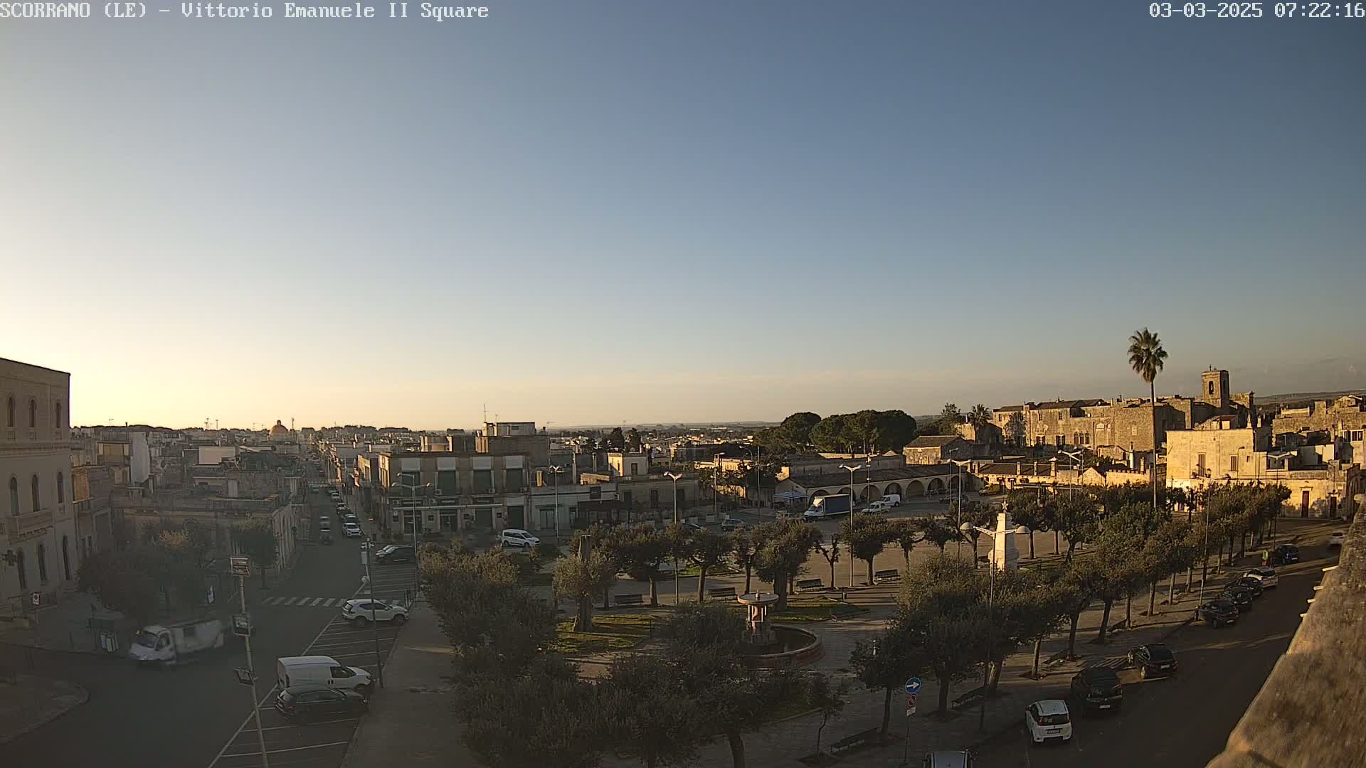 A clear, twilight view of a town square, with buildings, trees, parked cars, and a fountain, under a mostly clear sky.