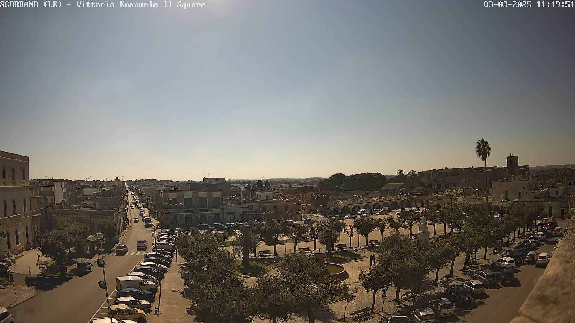 A sunny day reveals a town square with parked cars,  trees, and a street leading to a distant city under a clear sky.