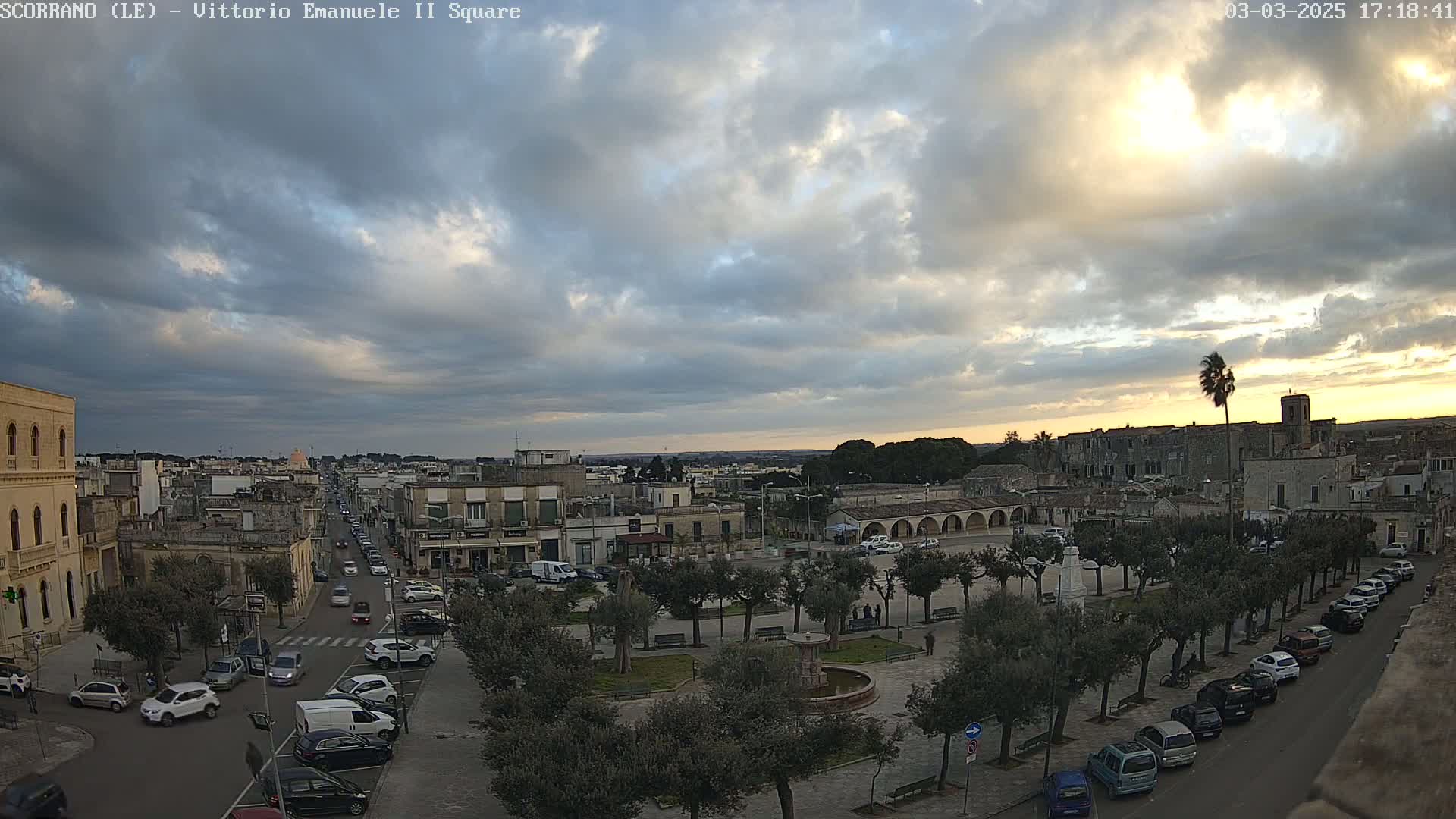 A partly cloudy sunset illuminates a town square with cars parked along the edges and a fountain in the center, surrounded by trees.