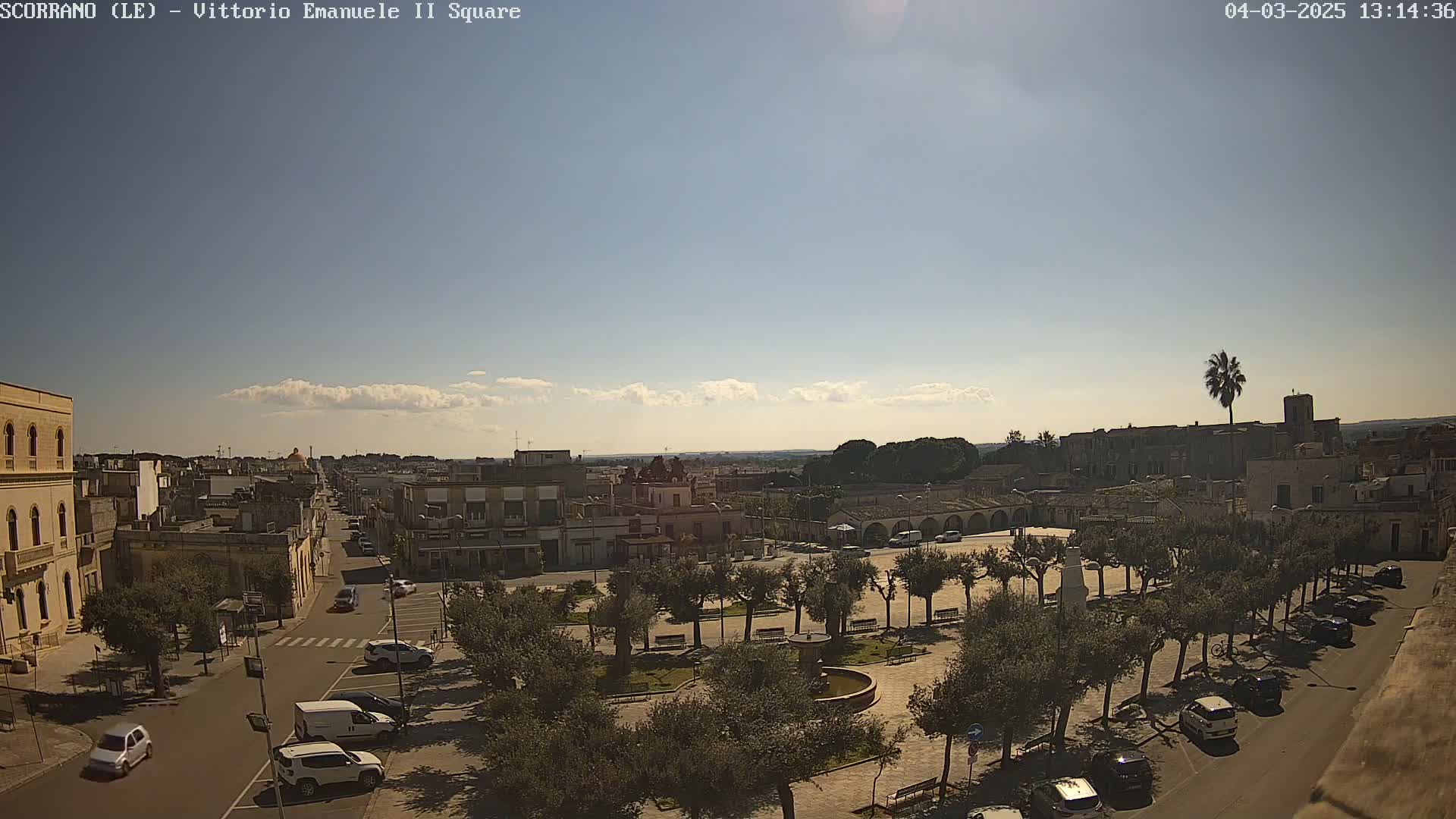 A sunny day reveals a town square with parked cars, trees lining the streets, and buildings extending into the distance under a mostly clear sky.