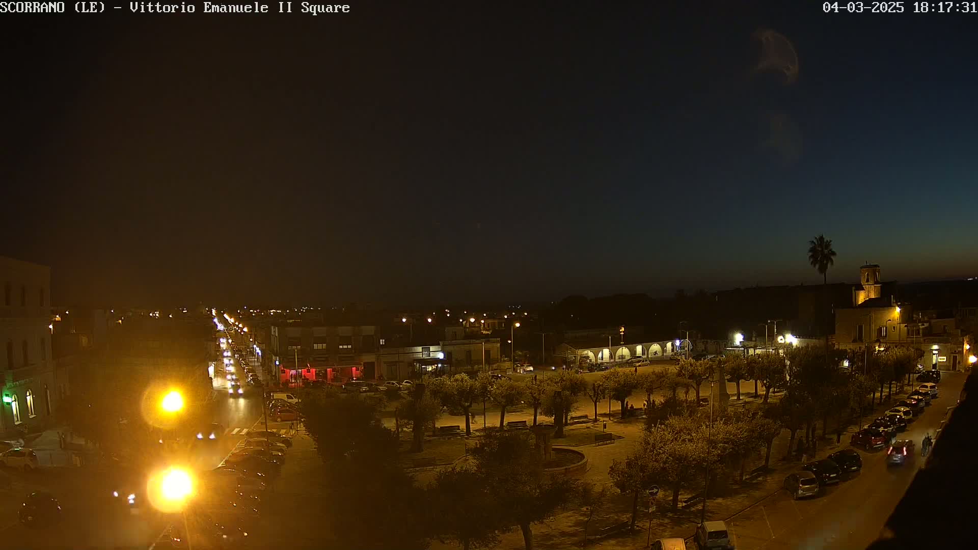 A nighttime view of a town square with trees, buildings, and cars, under a dark sky.