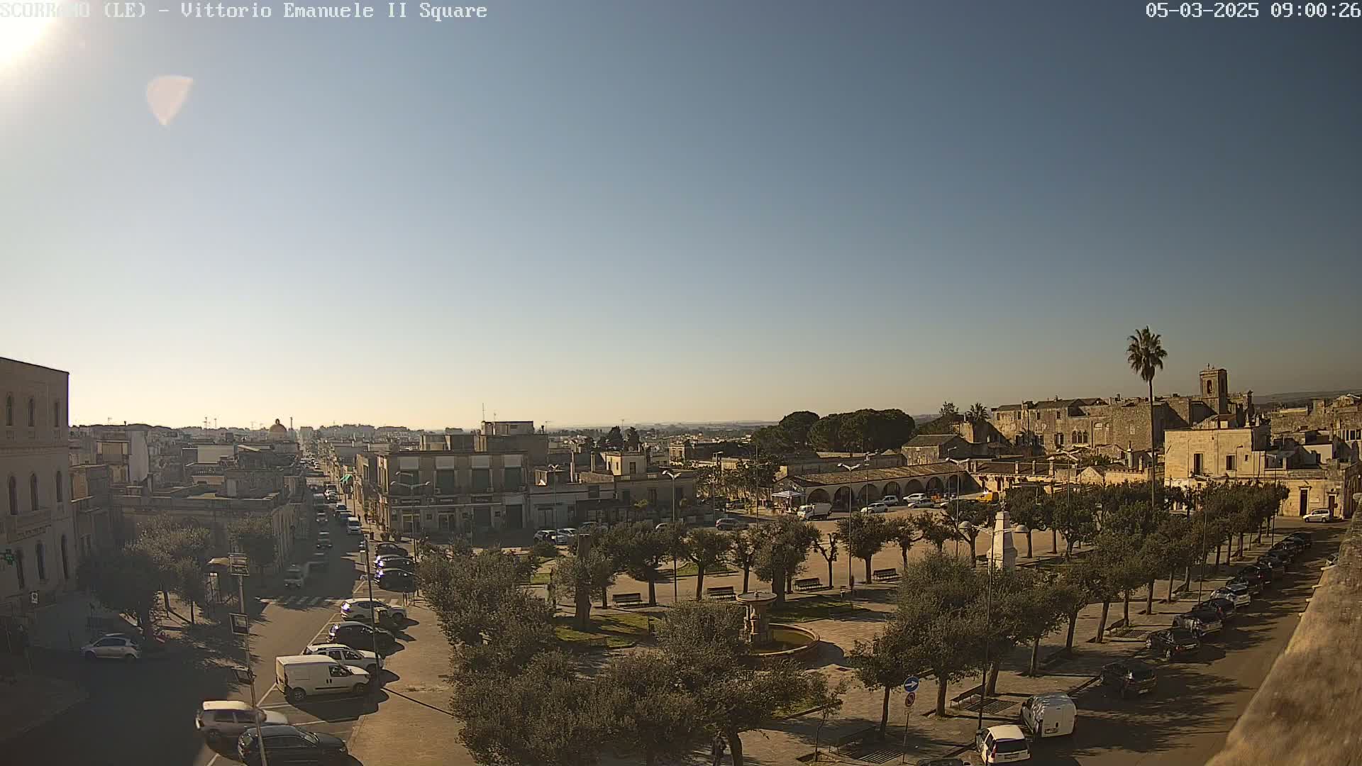 A clear, sunny day reveals a town square with a fountain, surrounded by olive trees and parked cars, leading to a street with more vehicles and buildings extending into the distance.