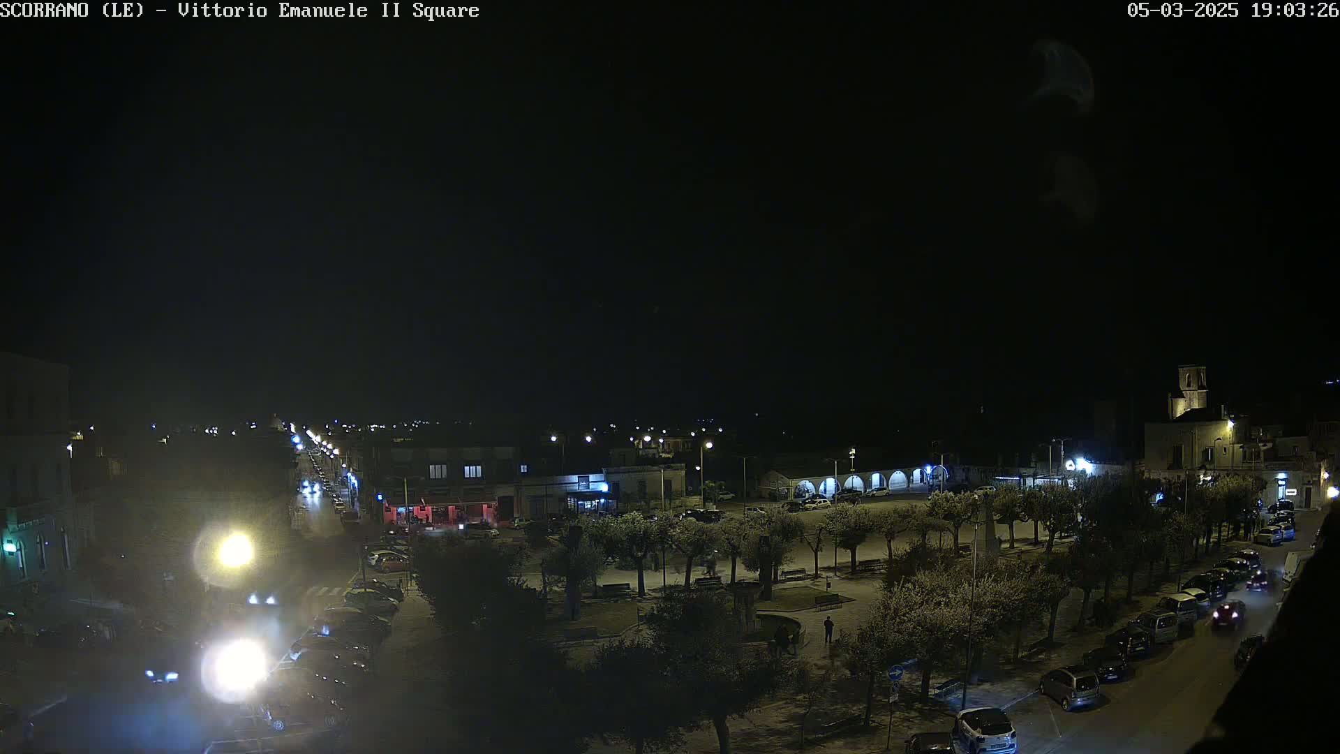 A nighttime, overhead view of a town square with parked cars lining the streets and a few pedestrians in the square.