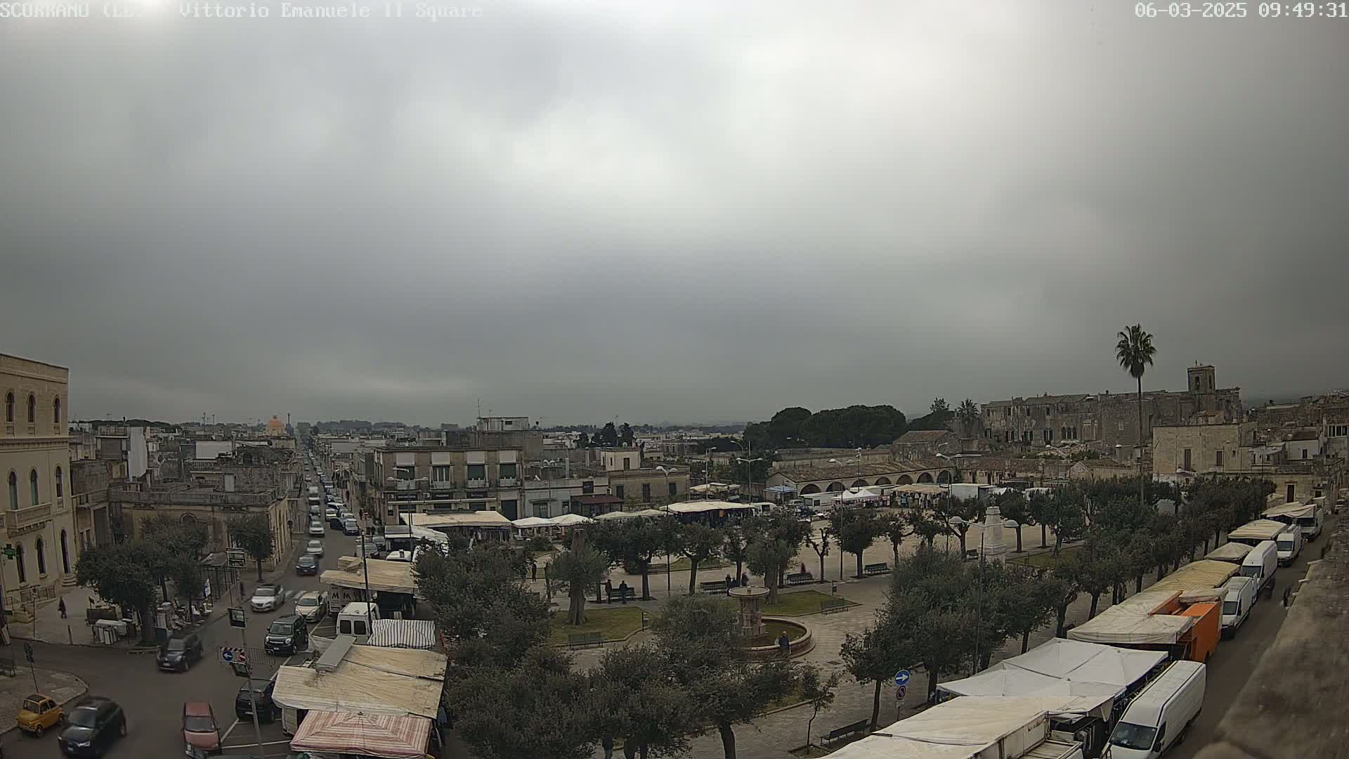 A town square with a market is shown under an overcast sky, with a street leading away from the square and buildings surrounding it.