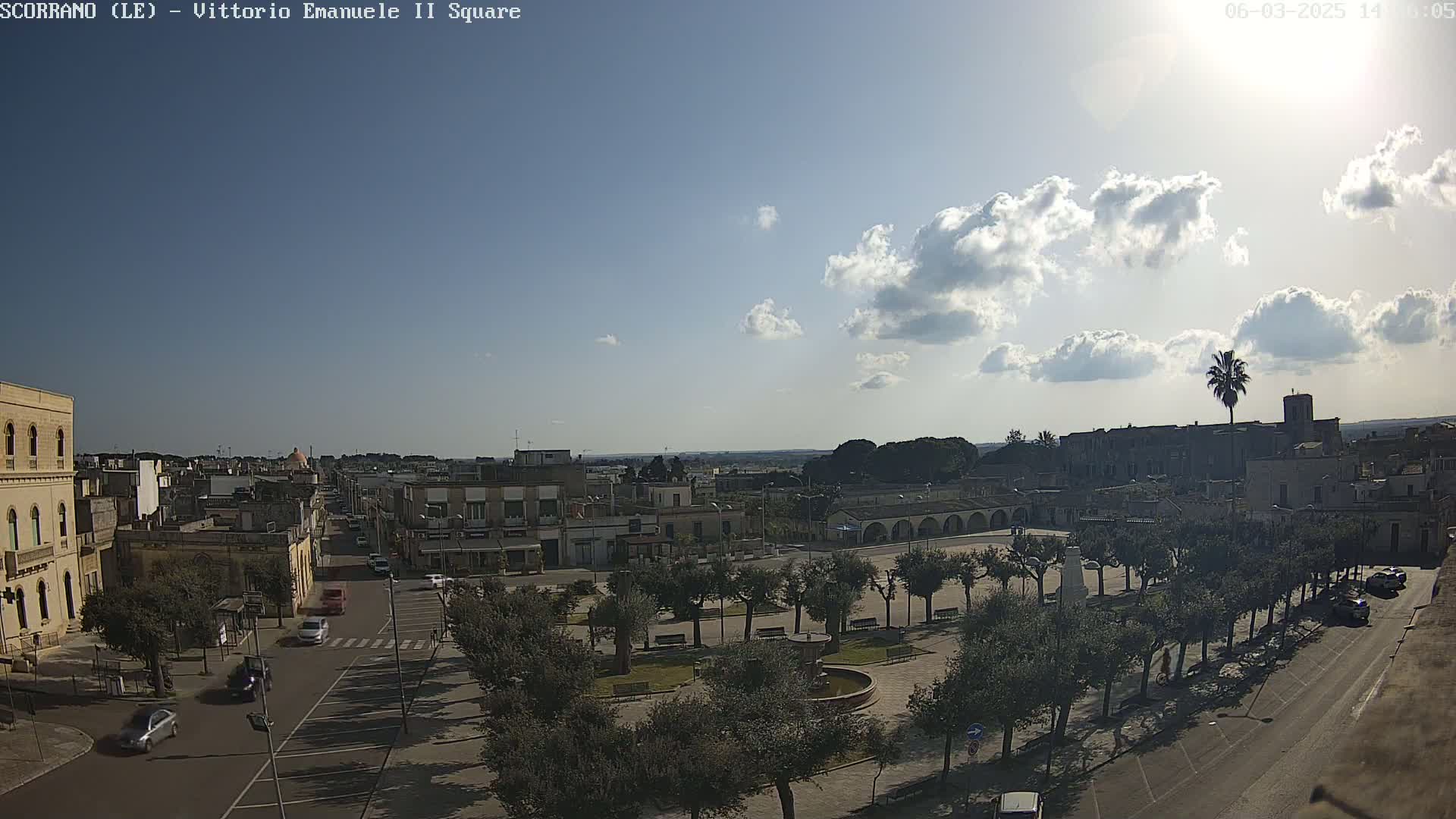 A partly sunny day reveals a town square with a fountain, surrounded by trees, buildings, and parked cars, with a few cars driving along roads.