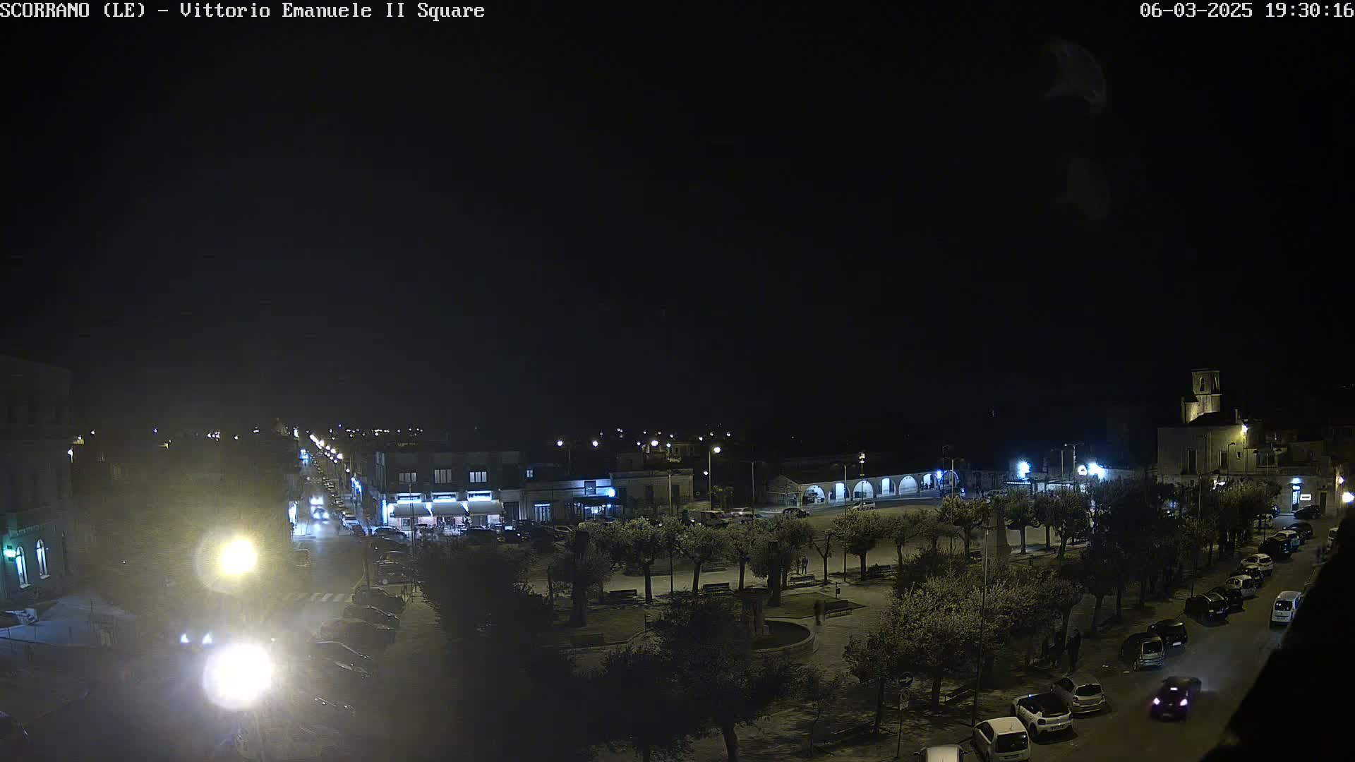 A nighttime view of a town square with parked cars and trees, illuminated by streetlights under a clear, dark sky.