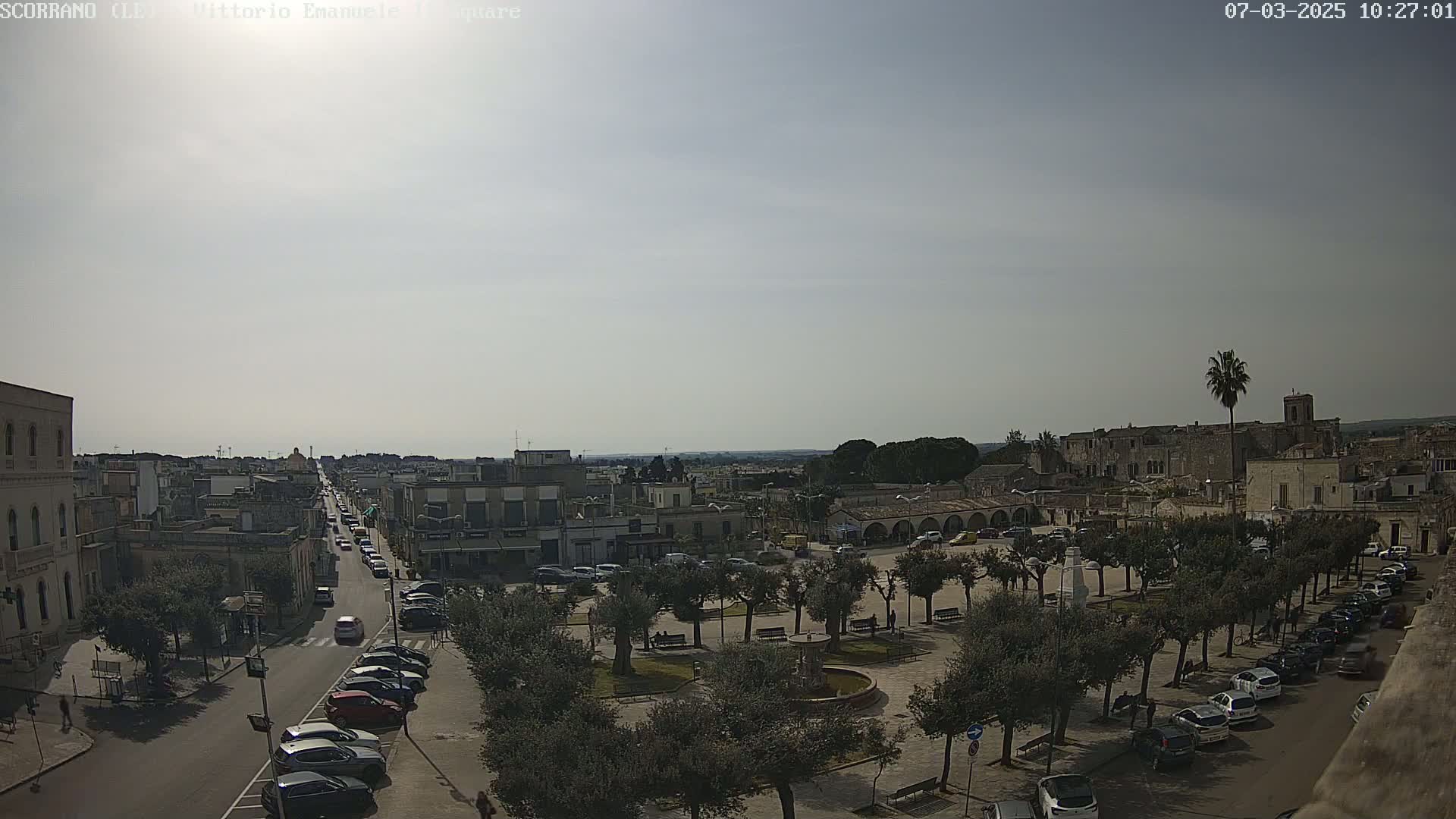 A sunny day reveals a town square with parked cars lining the streets, a central fountain, and tree-lined walkways, all overlooked by buildings and a distant, flat landscape.