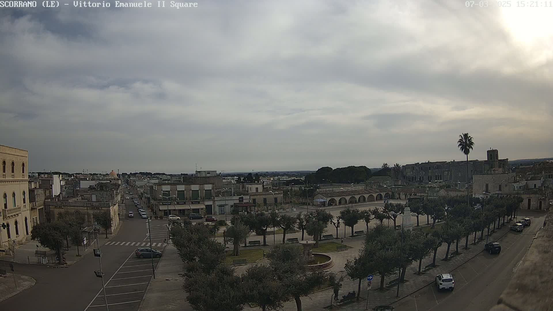 A town square with a fountain and rows of trees is visible under an overcast sky, with buildings and streets extending into the distance.