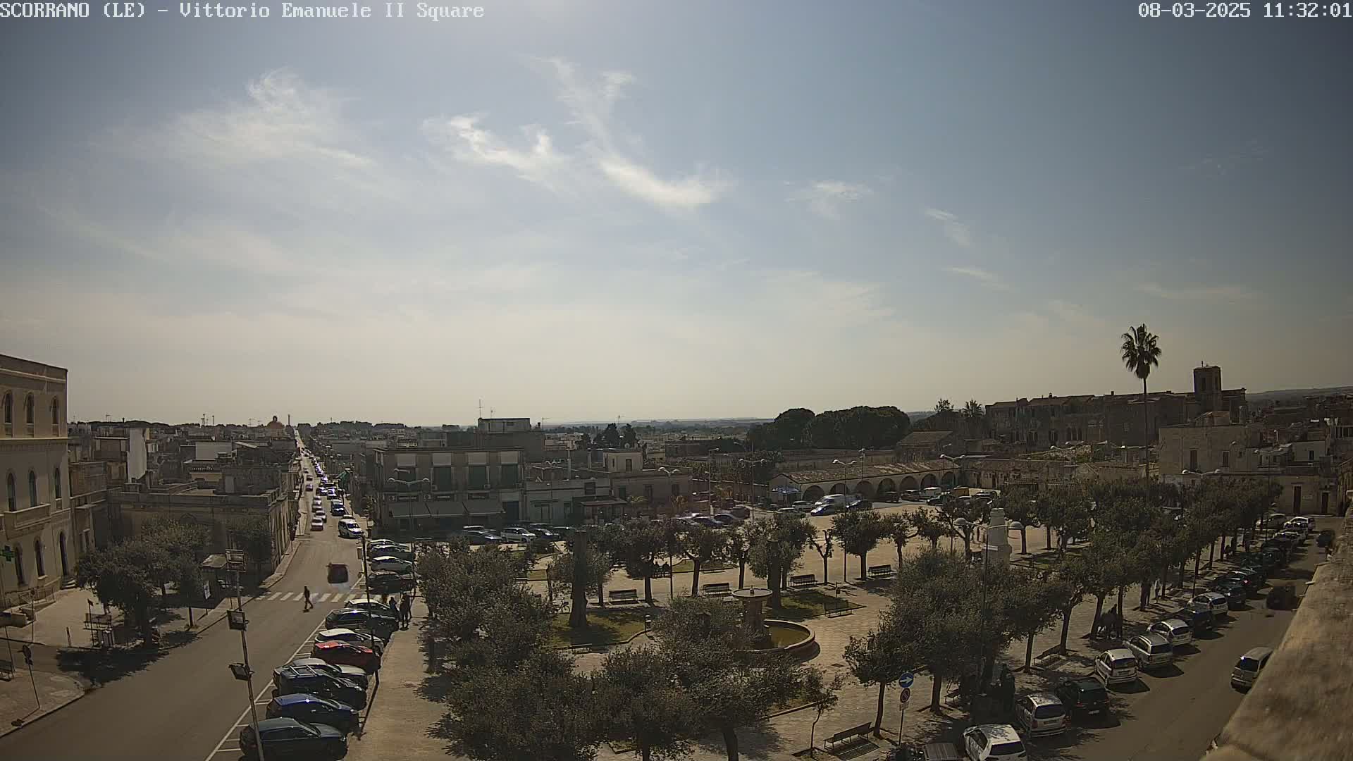 A mostly sunny day reveals a town square filled with parked cars and olive trees, with a street leading to a distant horizon.
