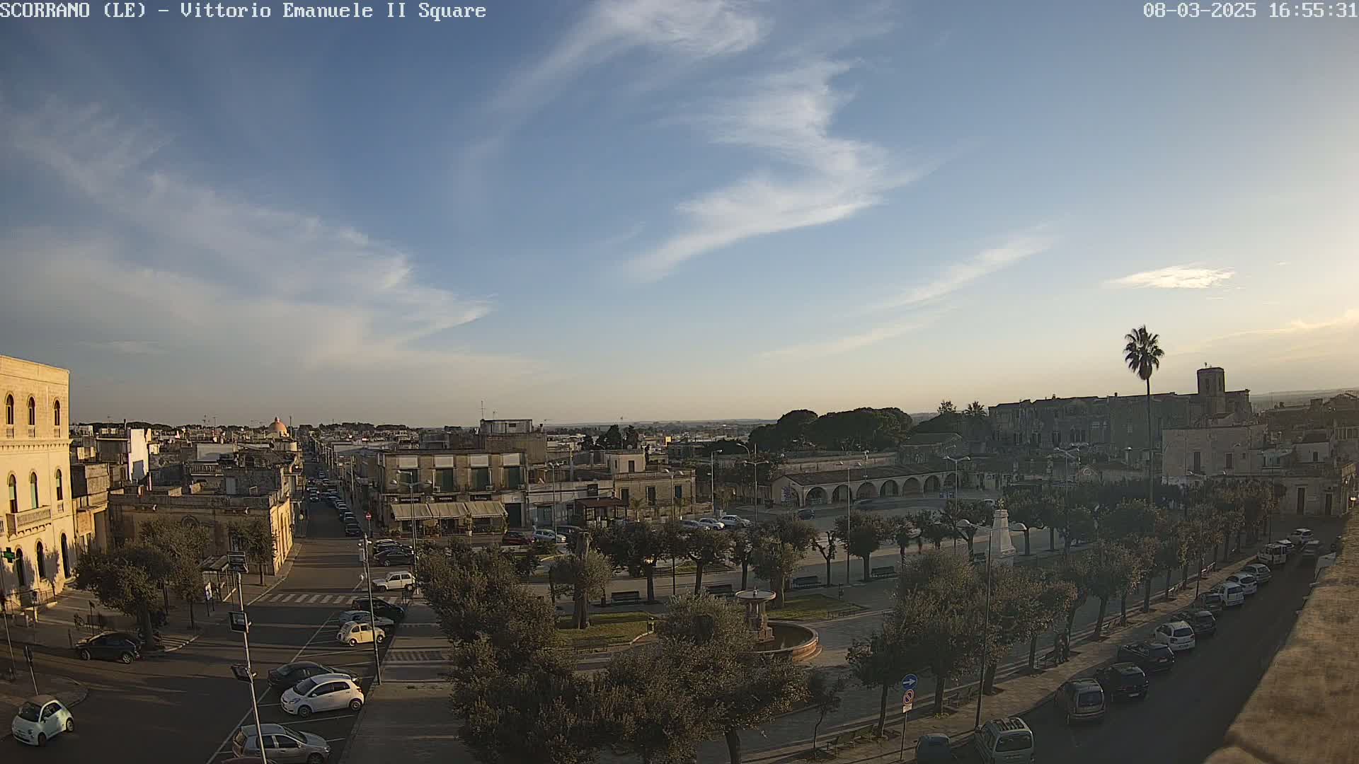 A mostly sunny, slightly cloudy day reveals a town square with parked cars, buildings, and trees, viewed from an elevated perspective.