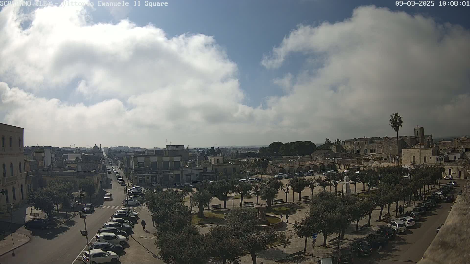 A partly cloudy day reveals a town square with a fountain, surrounded by parked cars and olive trees, with a street leading to more buildings in the distance.