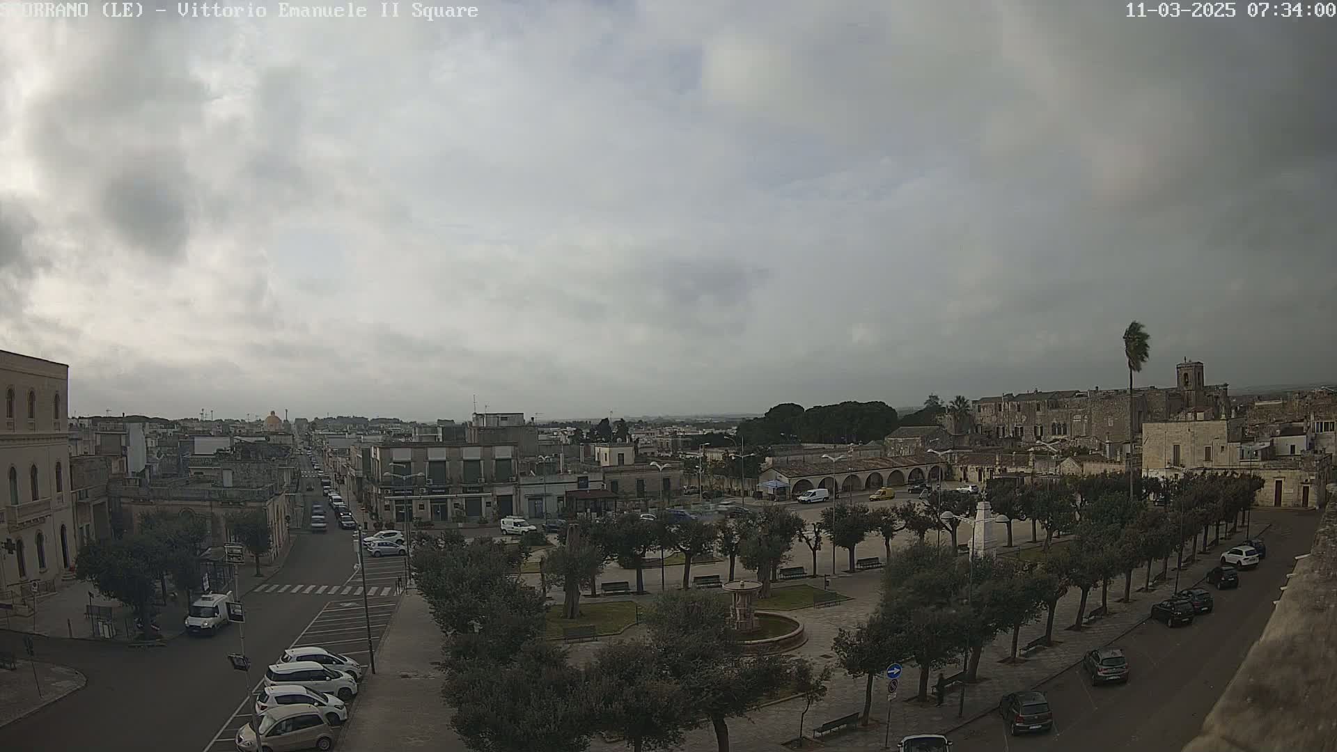 A town square with a fountain and parked cars is seen under an overcast sky.