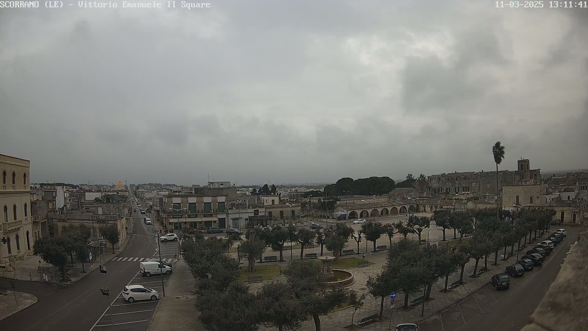 A town square with parked cars and a fountain is seen under an overcast sky.