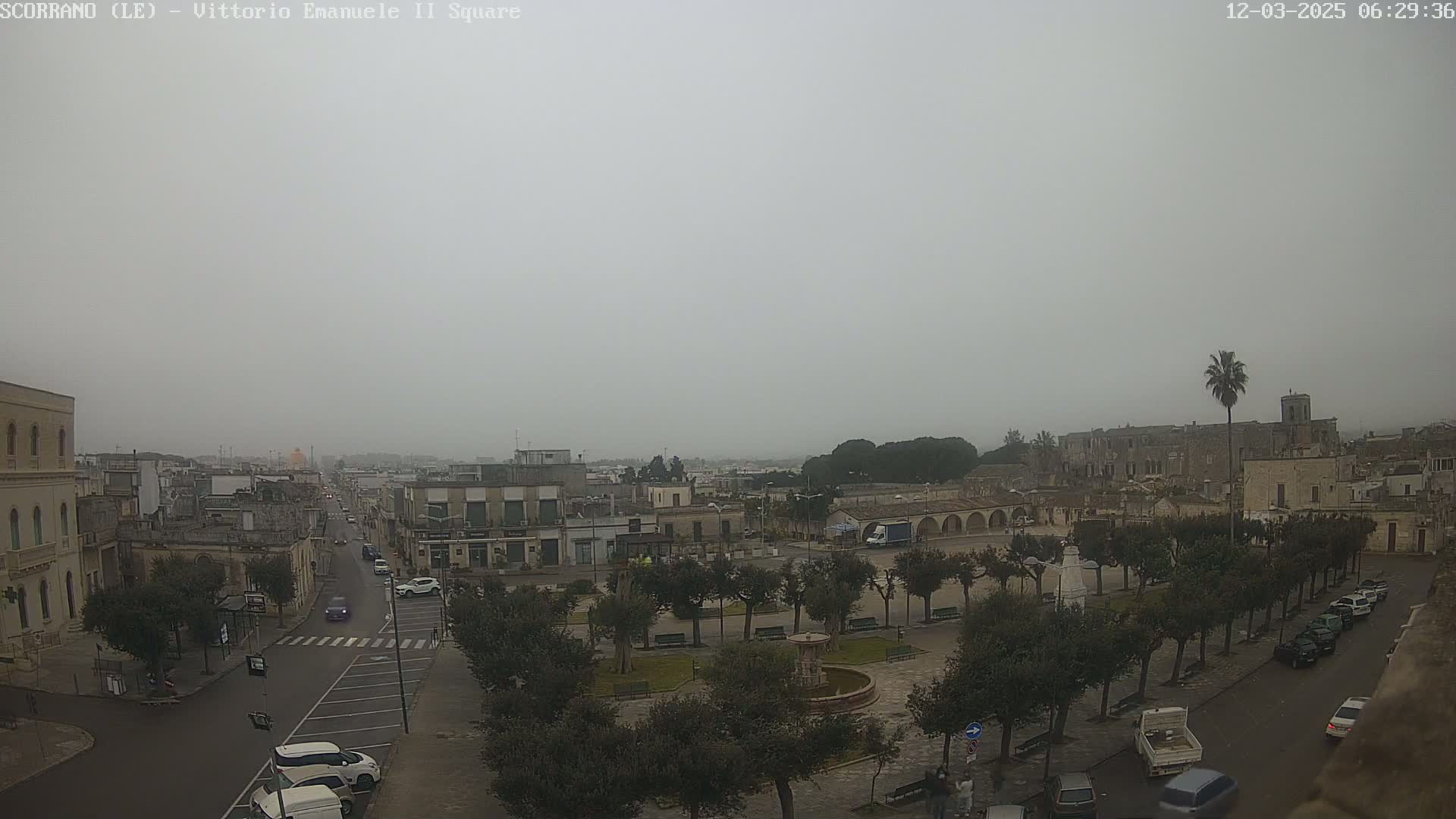 A foggy view of a town square with a fountain and surrounding buildings, cars parked along the streets, and several trees lining the square's perimeter.