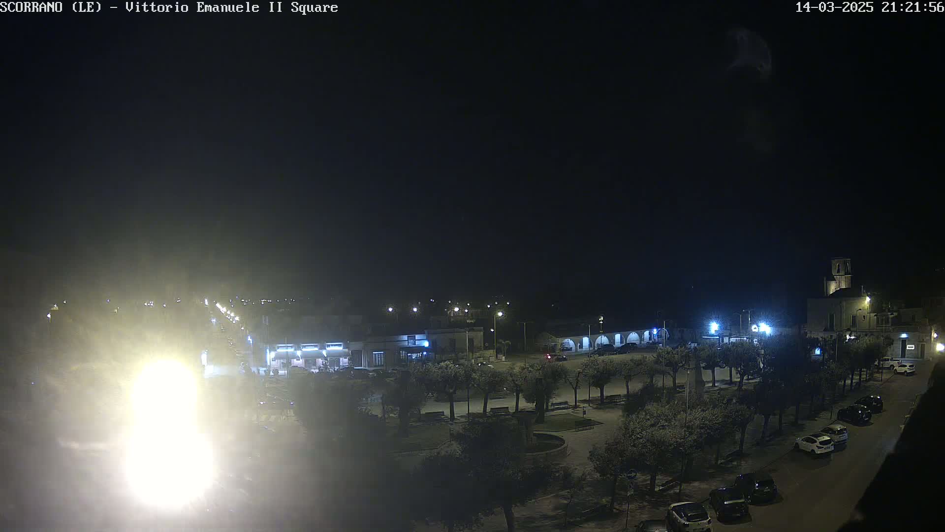 A nighttime view of a town square with cars parked along the edges, trees lining the perimeter, and buildings illuminated by streetlights.